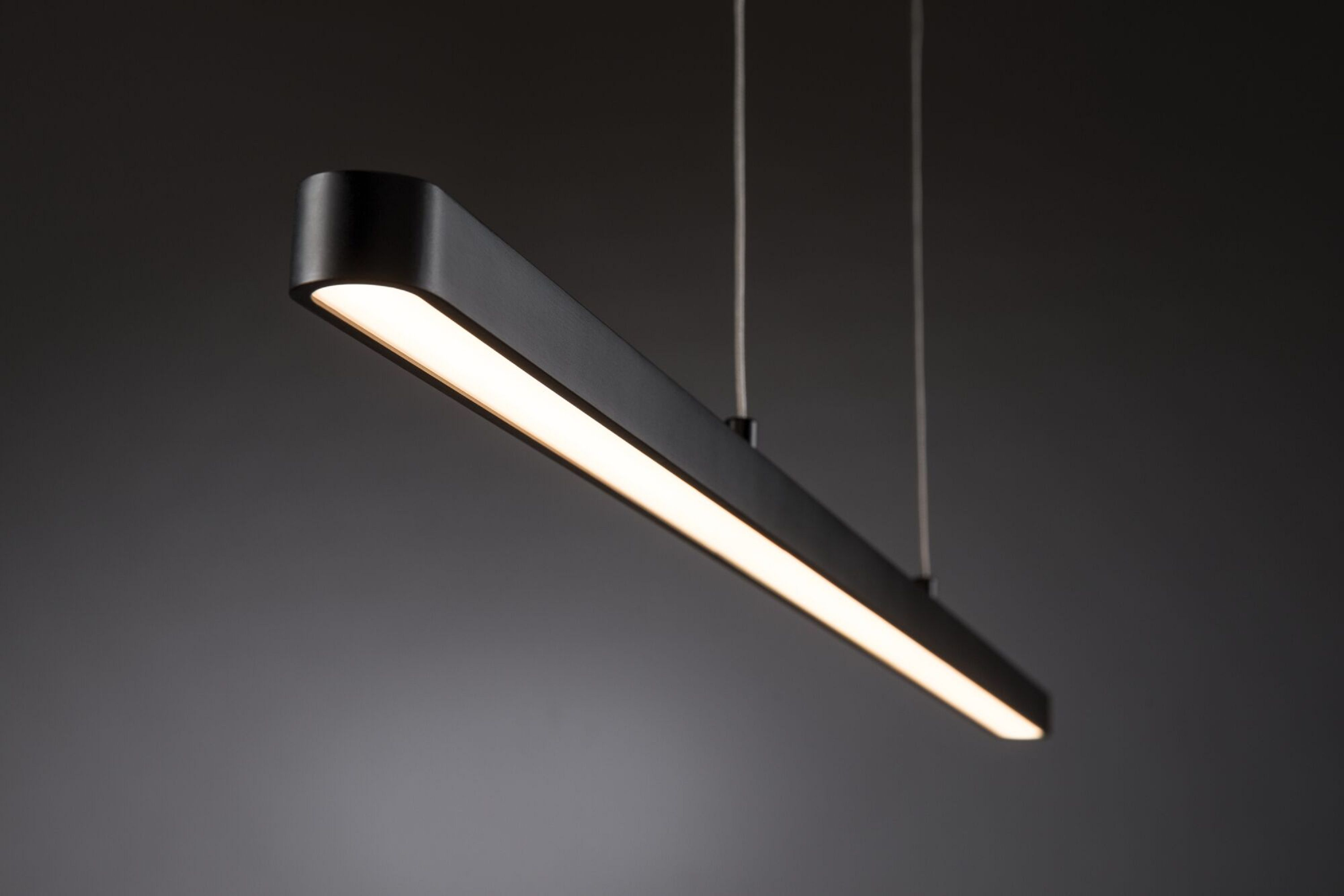Czarna prostokątna lampa wisząca z jasnym białym paskiem świetlnym, wisząca na cienkich przewodach, na szarym tle.