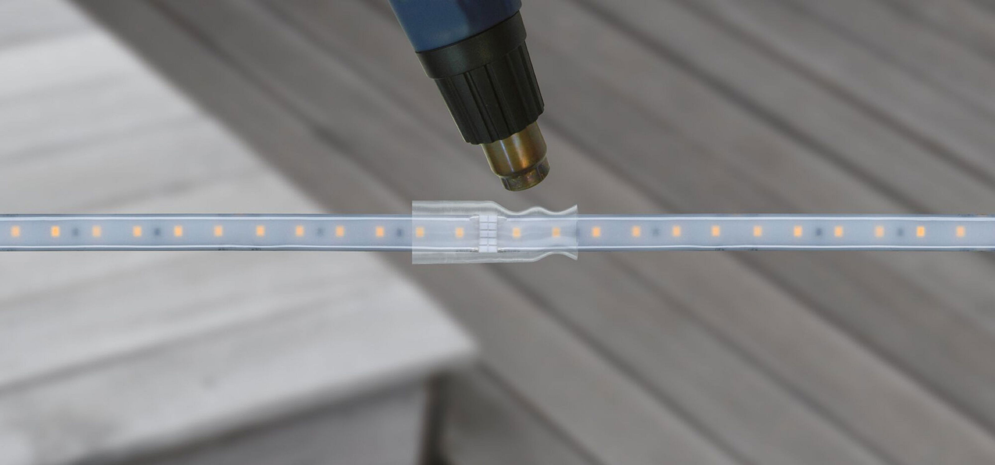 Eine Heißluftpistole zeigt auf einen transparenten Kunststoffverbinder auf einem Lichtband, möglicherweise zum Abdichten. Hintergrund sind Holzplanken.