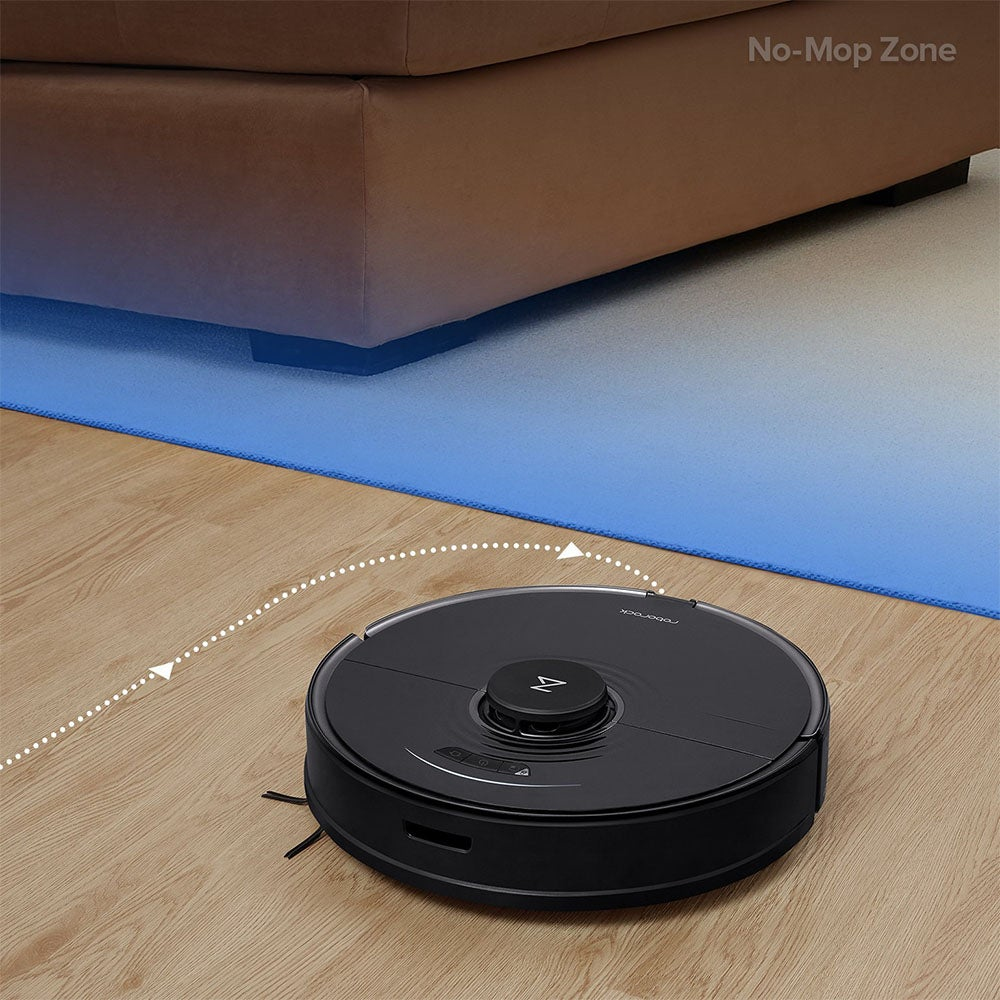 Ein schwarzer Roboter-Staubsauger auf einem Holzboden. Im Hintergrund ein braunes Sofa. In der Mitte liegt eine blaue Matte.