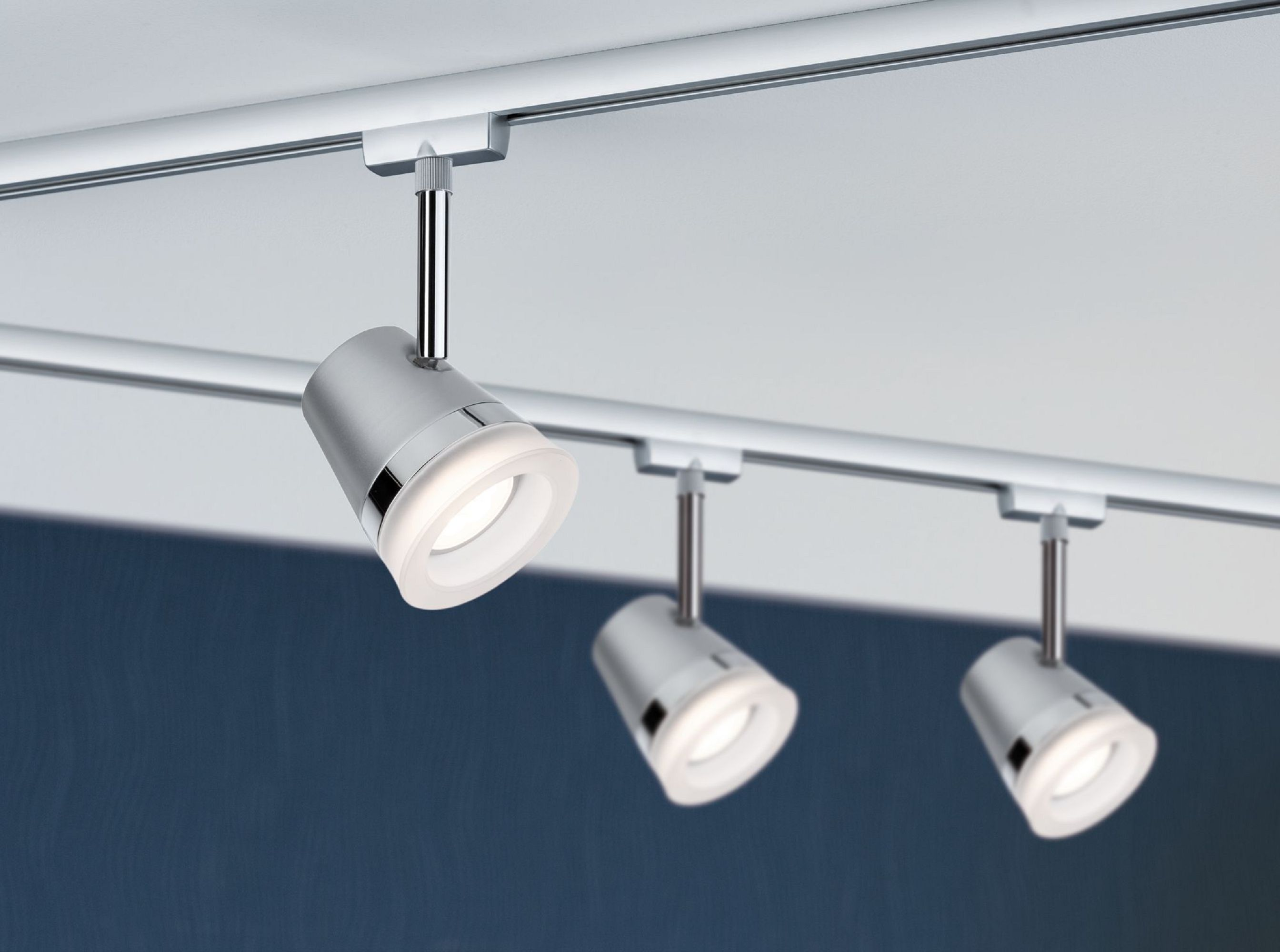 Tre faretti argentati su un soffitto bianco. Ogni luce è angolata verso il basso, illuminando la superficie sottostante. Sfondo blu.