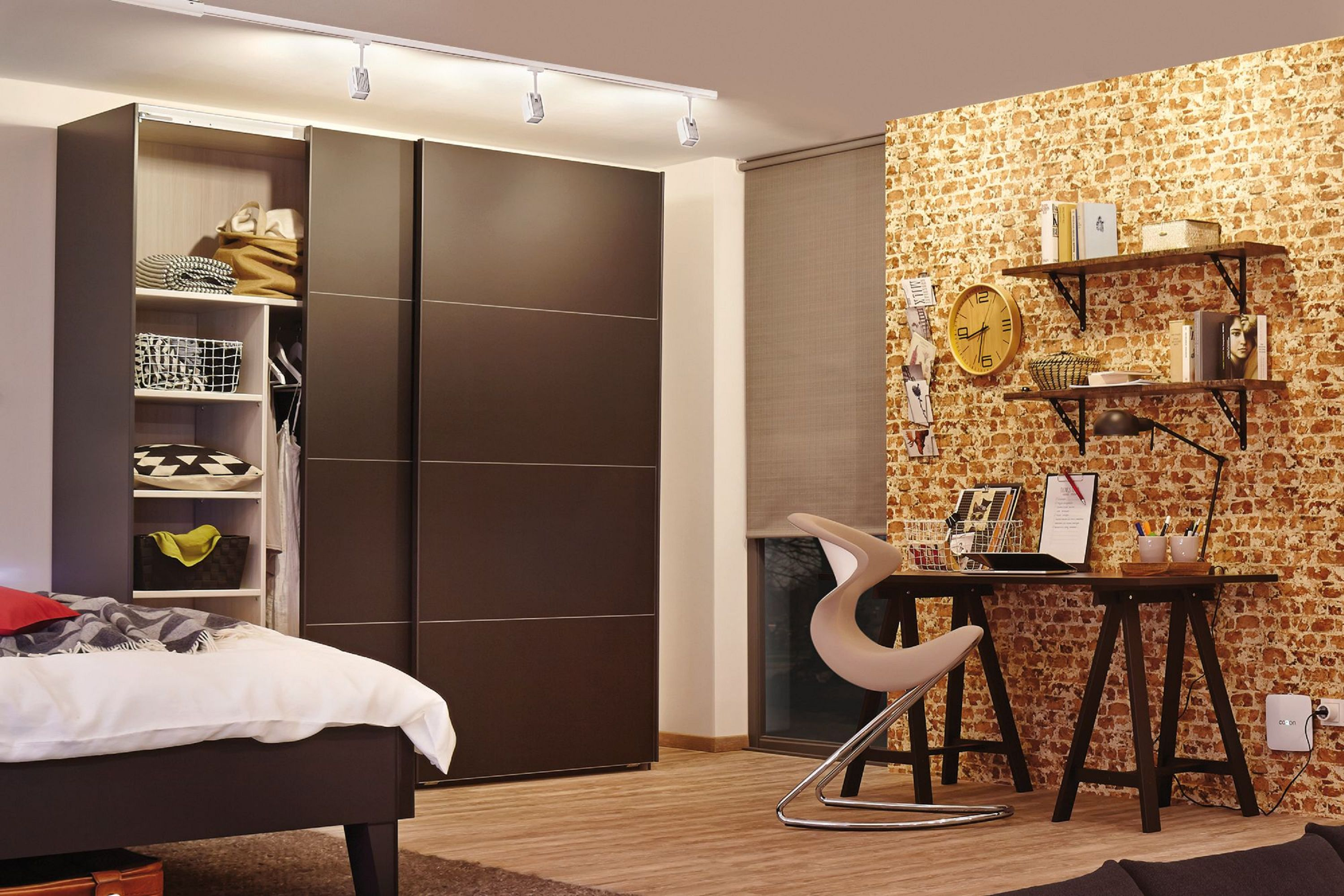 Schlafzimmer mit Schrank, Bett und Schreibtisch. Eine Ziegelmauer hat Regale mit Büchern und eine Uhr. Ein moderner Stuhl steht am Schreibtisch.
