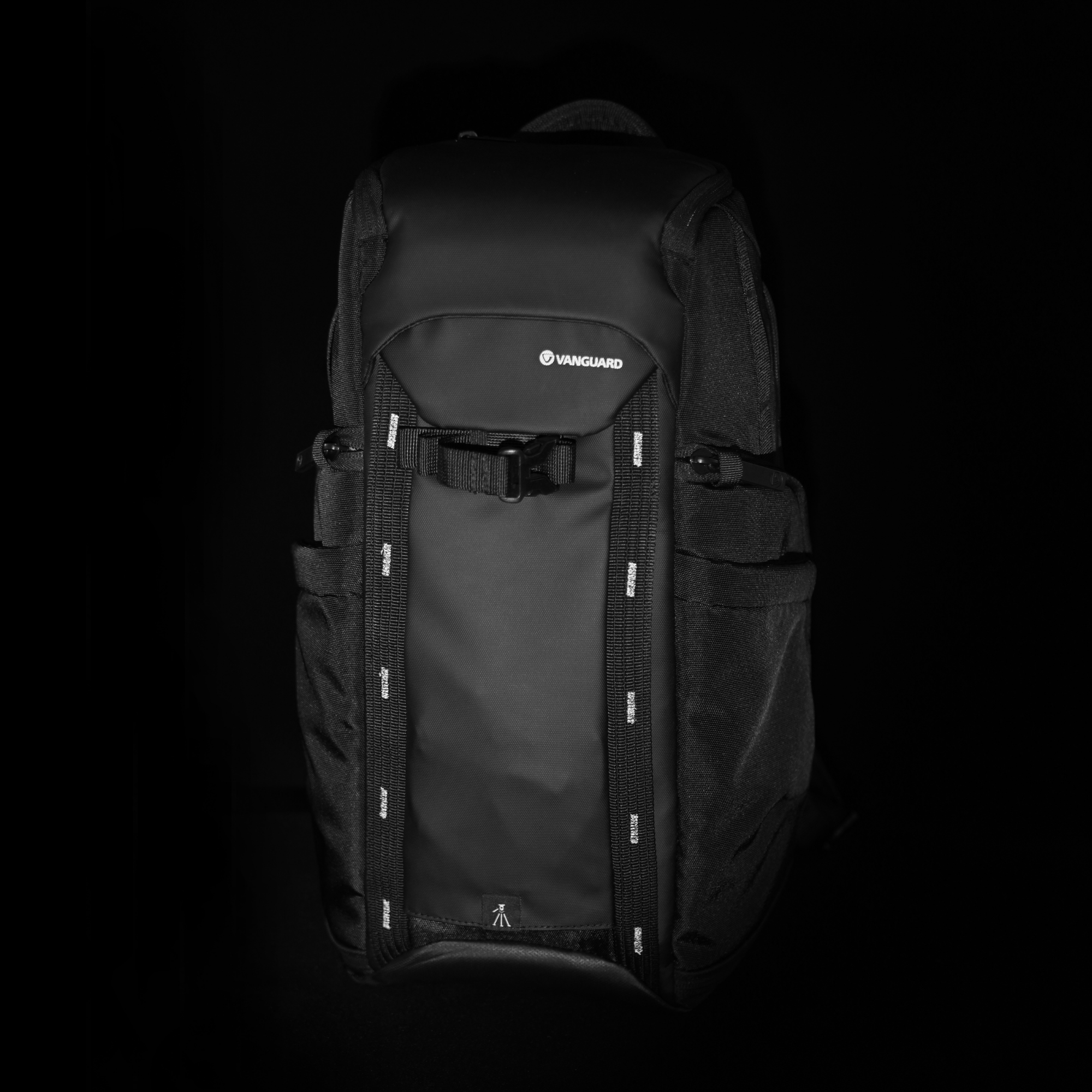 Mochila negra con el logo de Vanguard. Cuenta con correas, hebillas y bolsillos. Sobre un fondo oscuro, en escala de grises.
