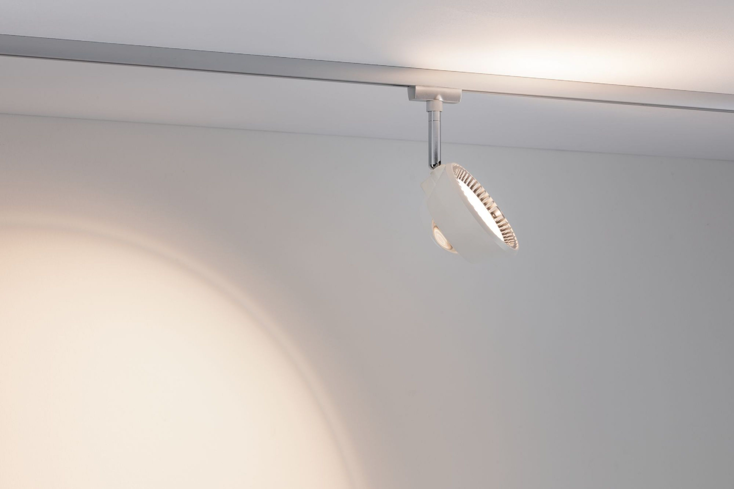 Ein weißer Scheinwerfer hängt an einer Schiene an einer weißen Decke und wirft einen kreisförmigen Lichtschein auf die Wand.