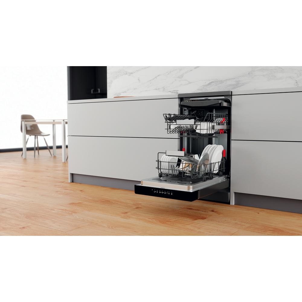 Otwarta zmywarka z naczyniami w środku. Drewniana podłoga, szare szafki i krzesło ze stołem w tle.