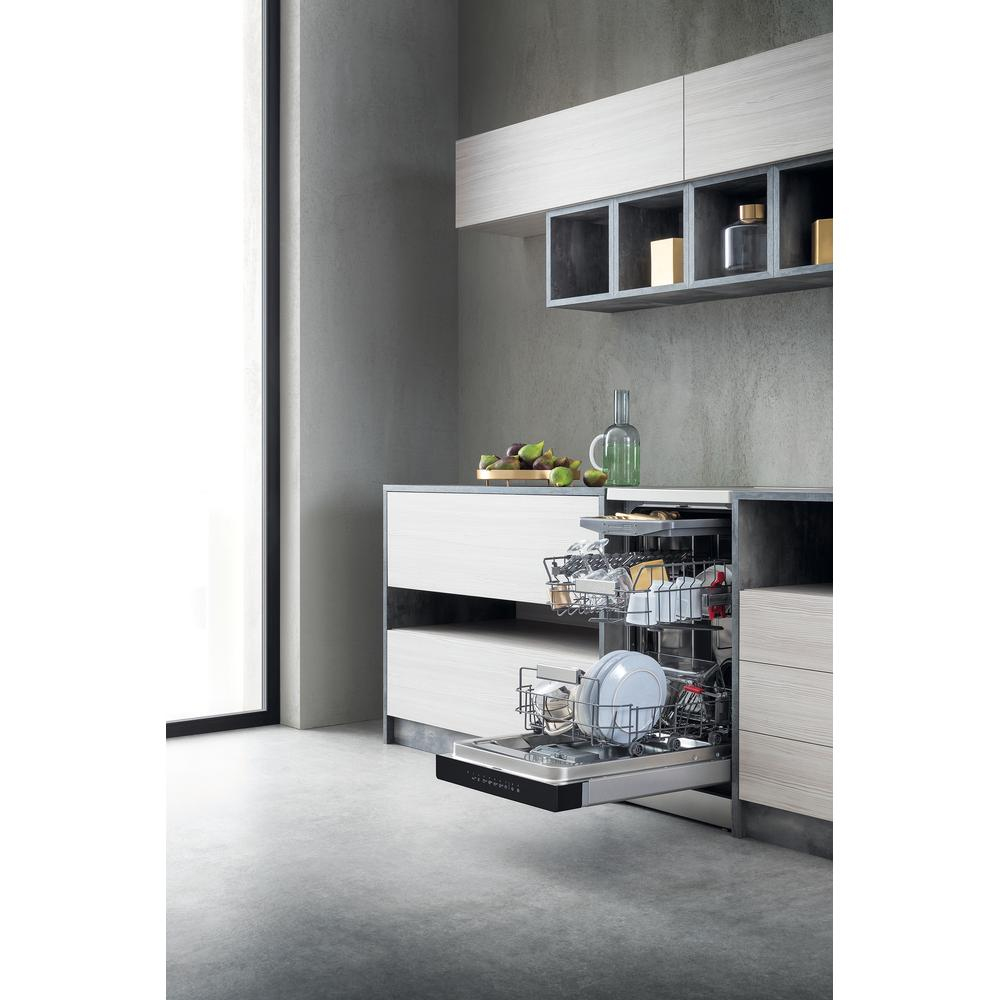 Scena kuchenna z otwartą zmywarką, załadowaną naczyniami. Widoczne są również szare szafki, owoce i szklana butelka.