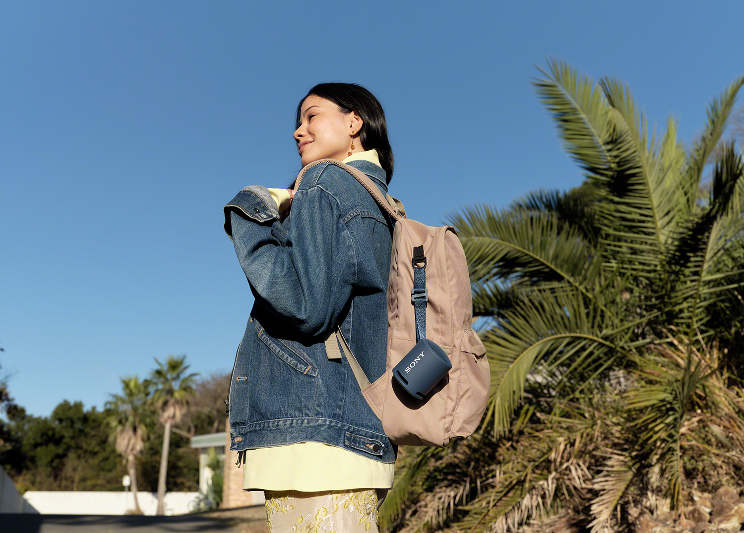 Donna con zaino e giacca di jeans, sorridente, tiene in mano un altoparlante Sony blu. Palme e cielo blu sullo sfondo.