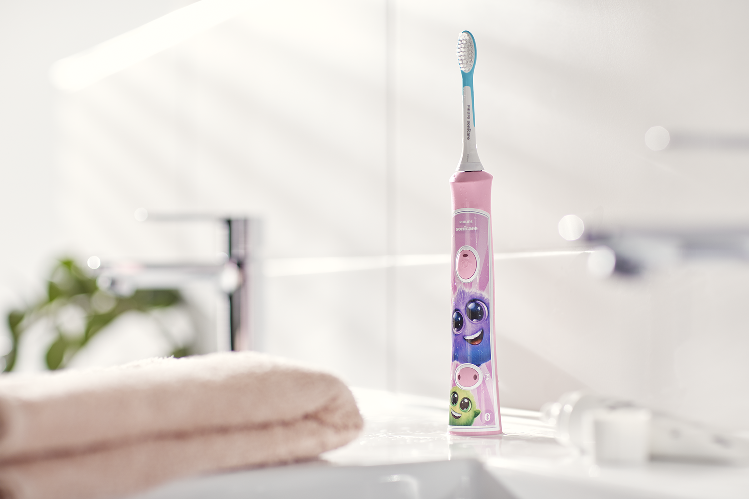 Un cepillo de dientes rosa con personajes de dibujos animados se encuentra sobre una superficie blanca. Una toalla beige y un baño borroso están al fondo.