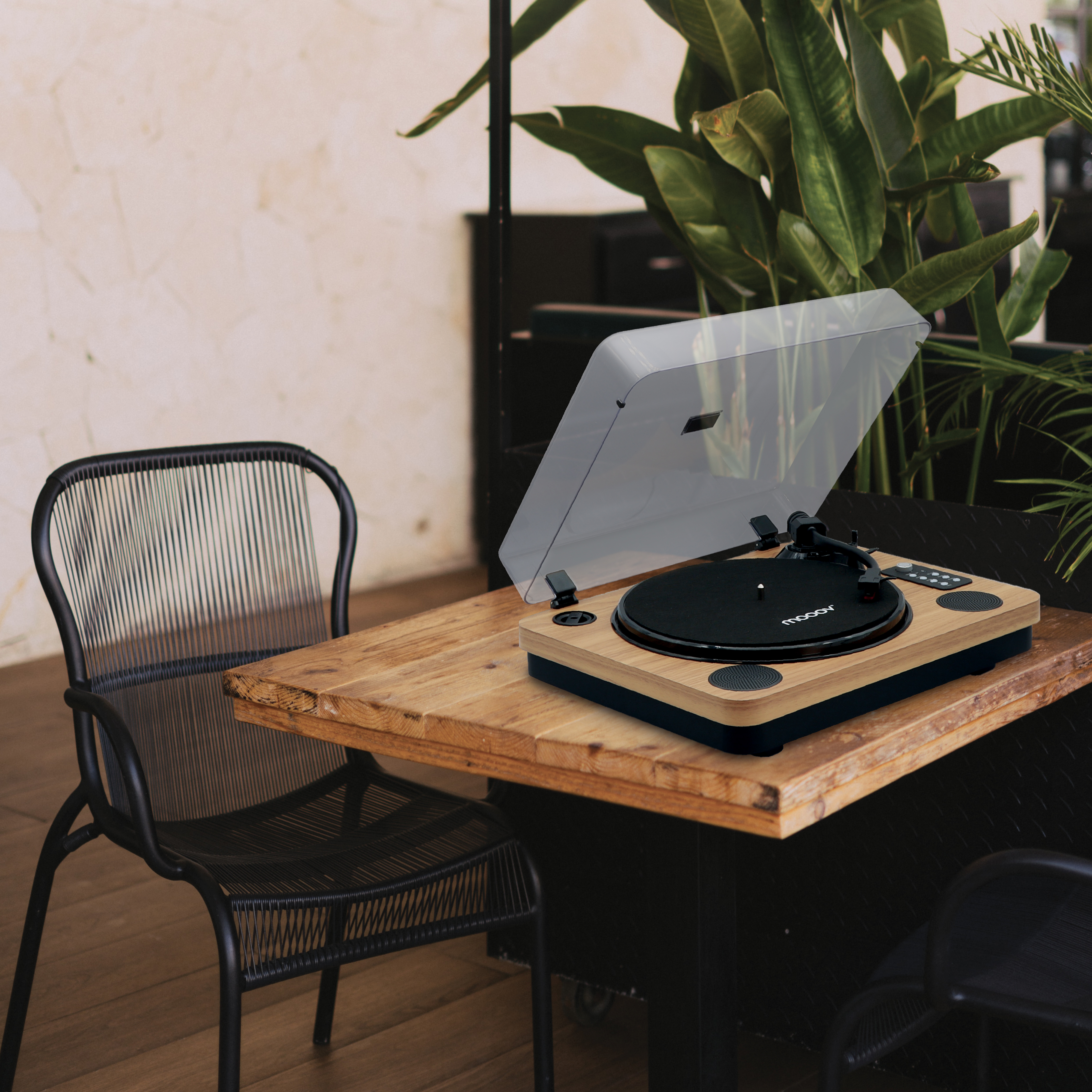 Un tocadiscos sobre una mesa de madera con una silla negra delante. Hojas verdes en el fondo.