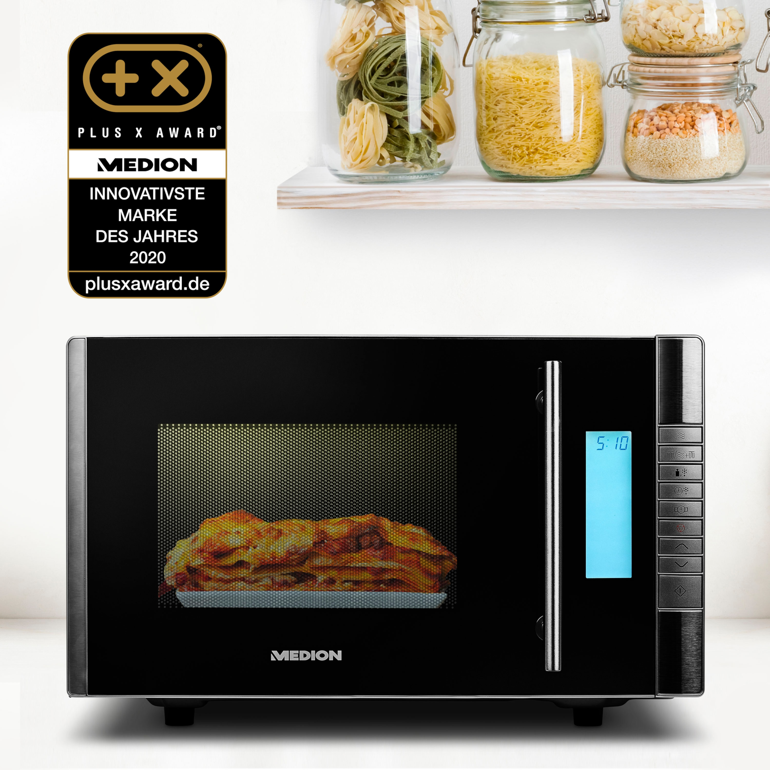 Un horno microondas negro con lasaña dentro. Tarros con pasta y granos en el fondo. Un premio plus x está a la izquierda.