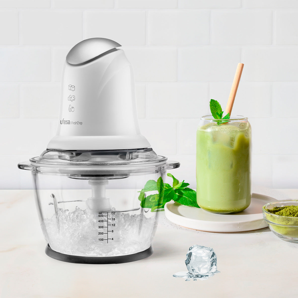 Un robot da cucina bianco con ghiaccio, una bevanda verde alla menta e una ciotolina di polvere verde.