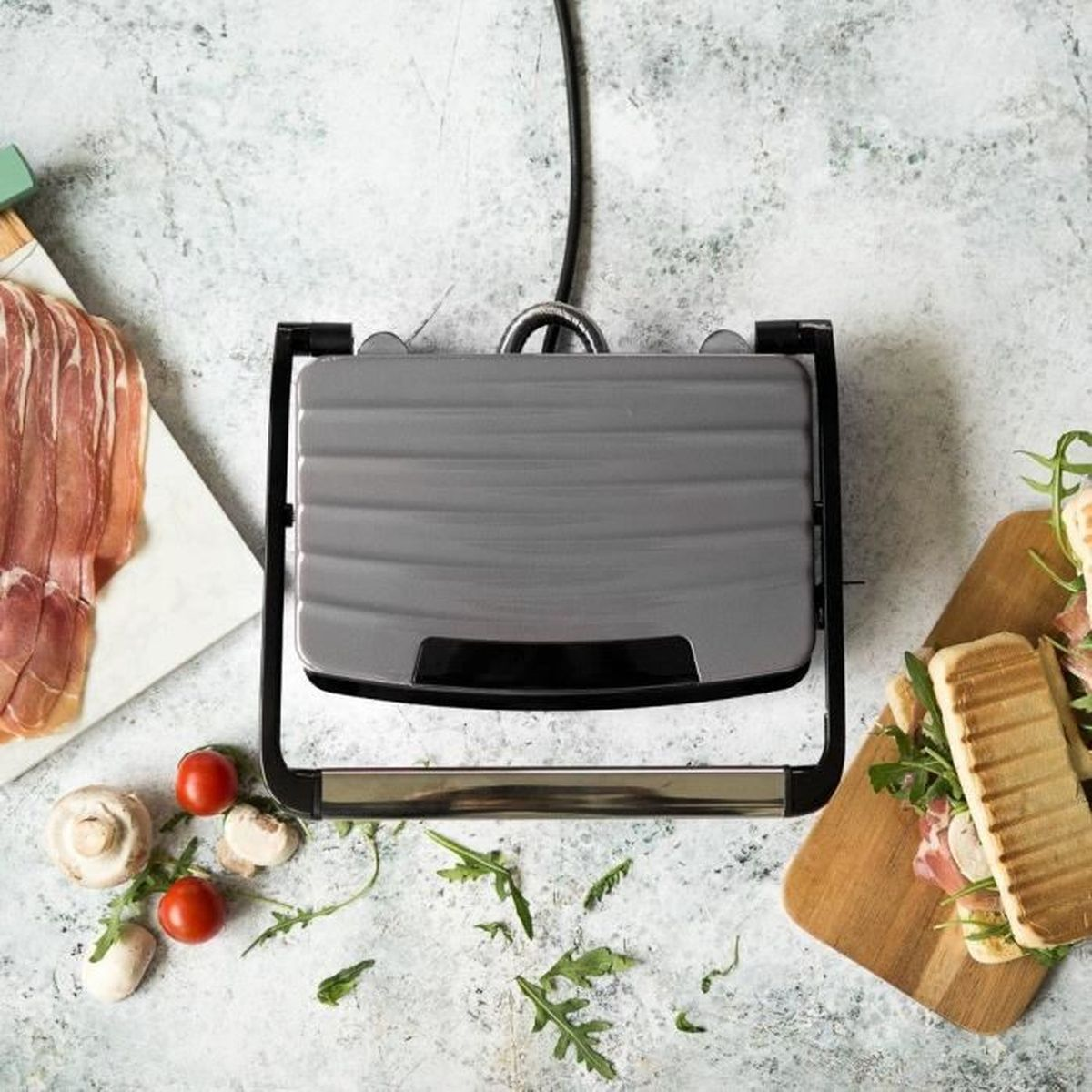 Una piastra per panini grigia con panini e ingredienti. Pancetta, pomodori, funghi e rucola sono disposti intorno alla piastra.