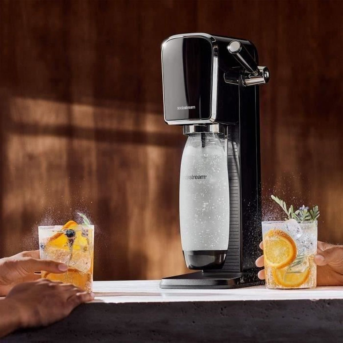Eine schwarze SodaStream-Maschine mit zwei Gläsern Cocktails. Eines hat Orangenscheiben und das andere Blaubeeren.