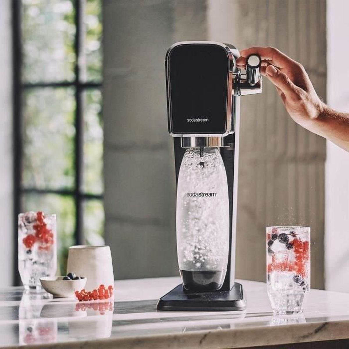 Eine SodaStream-Maschine, die von einer Hand bedient wird. Auf dem Tisch stehen zwei Gläser Wasser mit Beeren und eine Schüssel mit Beeren.