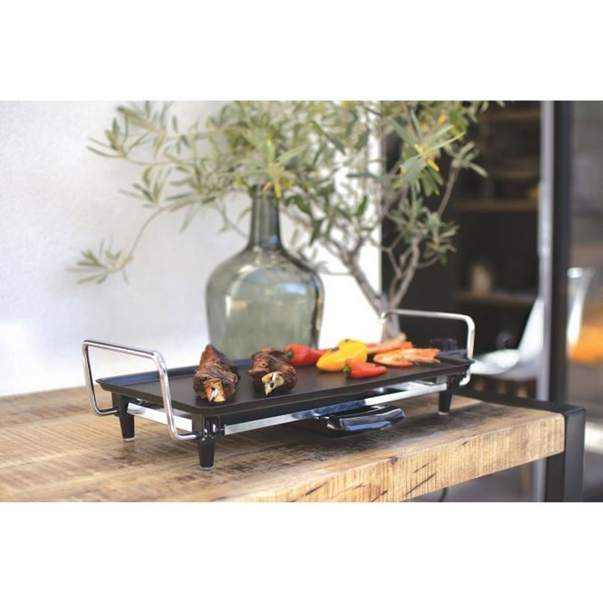 Una parrilla eléctrica negra con comida encima, colocada sobre una mesa de madera. Un jarrón con hojas verdes al fondo.