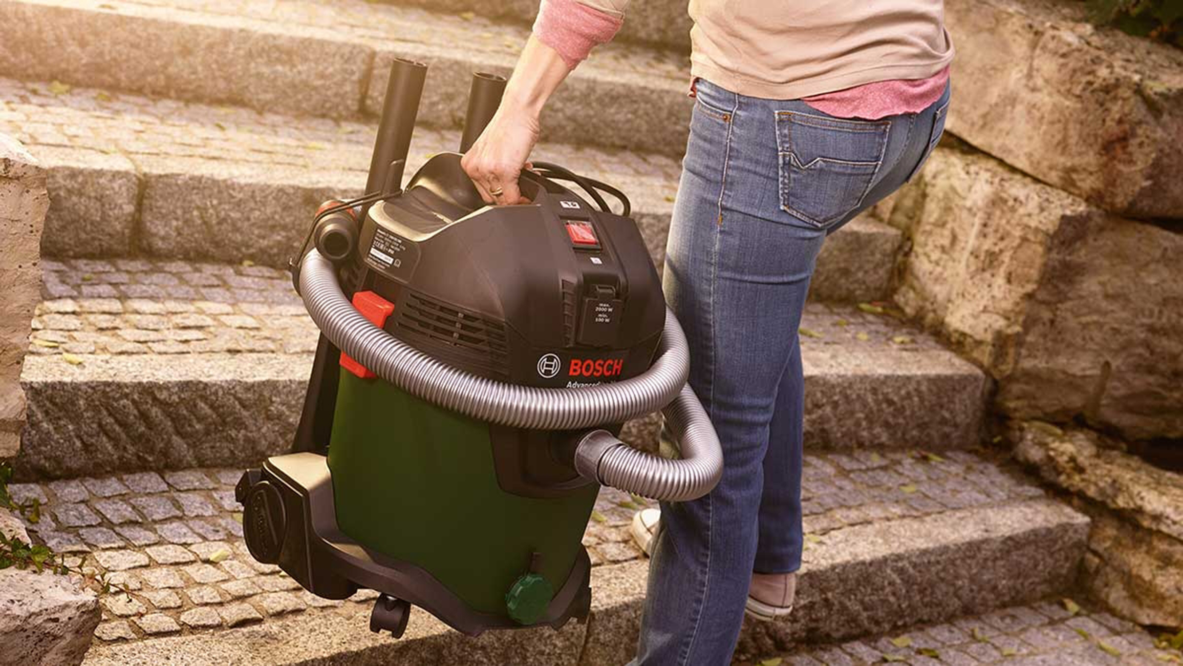 Bir kişi, Bosch marka bir elektrikli süpürgeyi taş basamaklardan yukarı taşıyor. Elektrikli süpürge siyah ve yeşil renktedir. Kişi kot pantolon ve pembe bir gömlek giyiyor.
