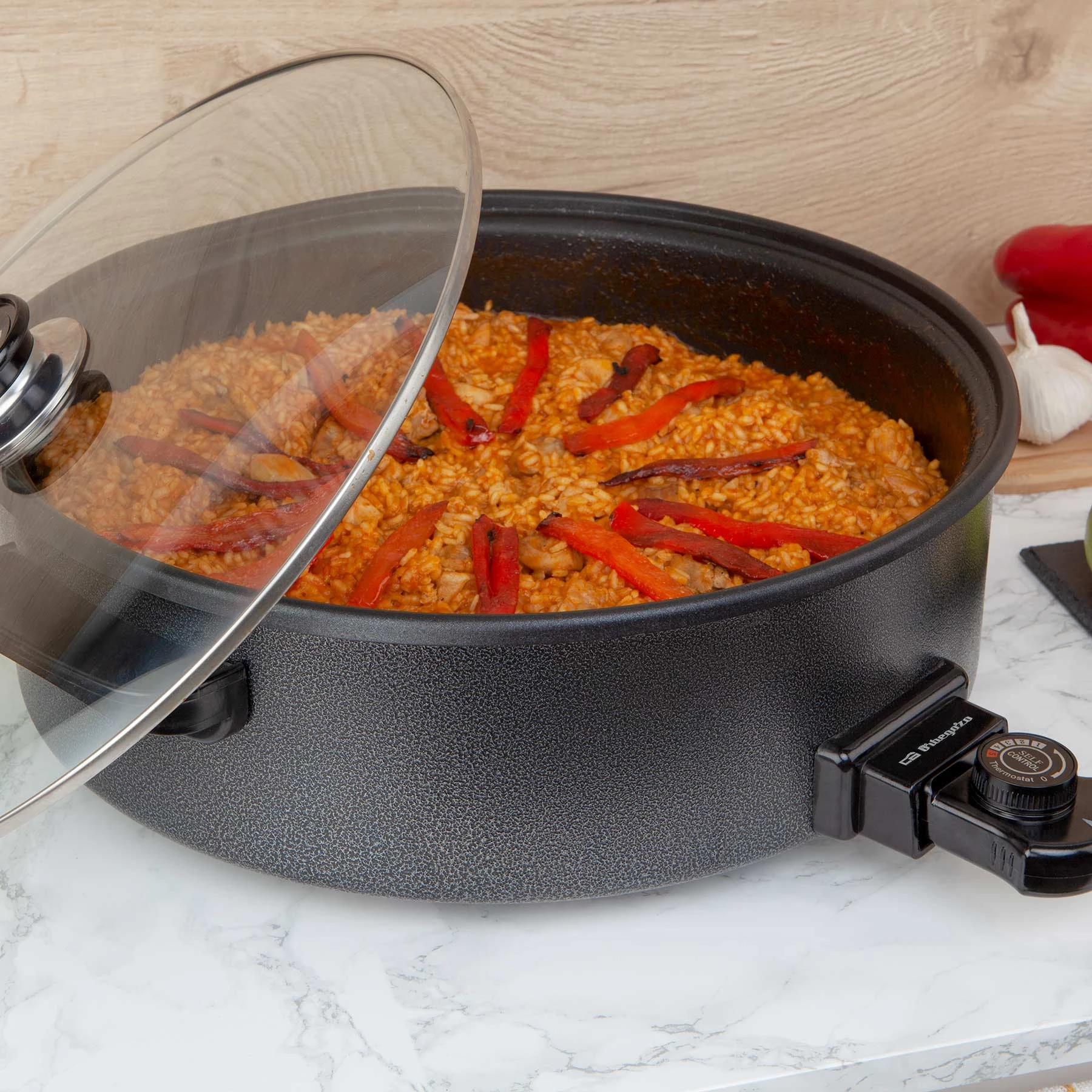 Paella en una sartén eléctrica negra con pimientos rojos. Tapa de cristal ligeramente abierta. Encimera de mármol y fondo de madera.