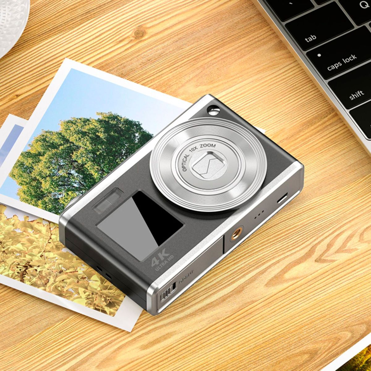 Aparat cyfrowy leży na drewnianej powierzchni obok zdjęć. Klawiatura laptopa jest widoczna w tle. Aparat jest srebrny i czarny.