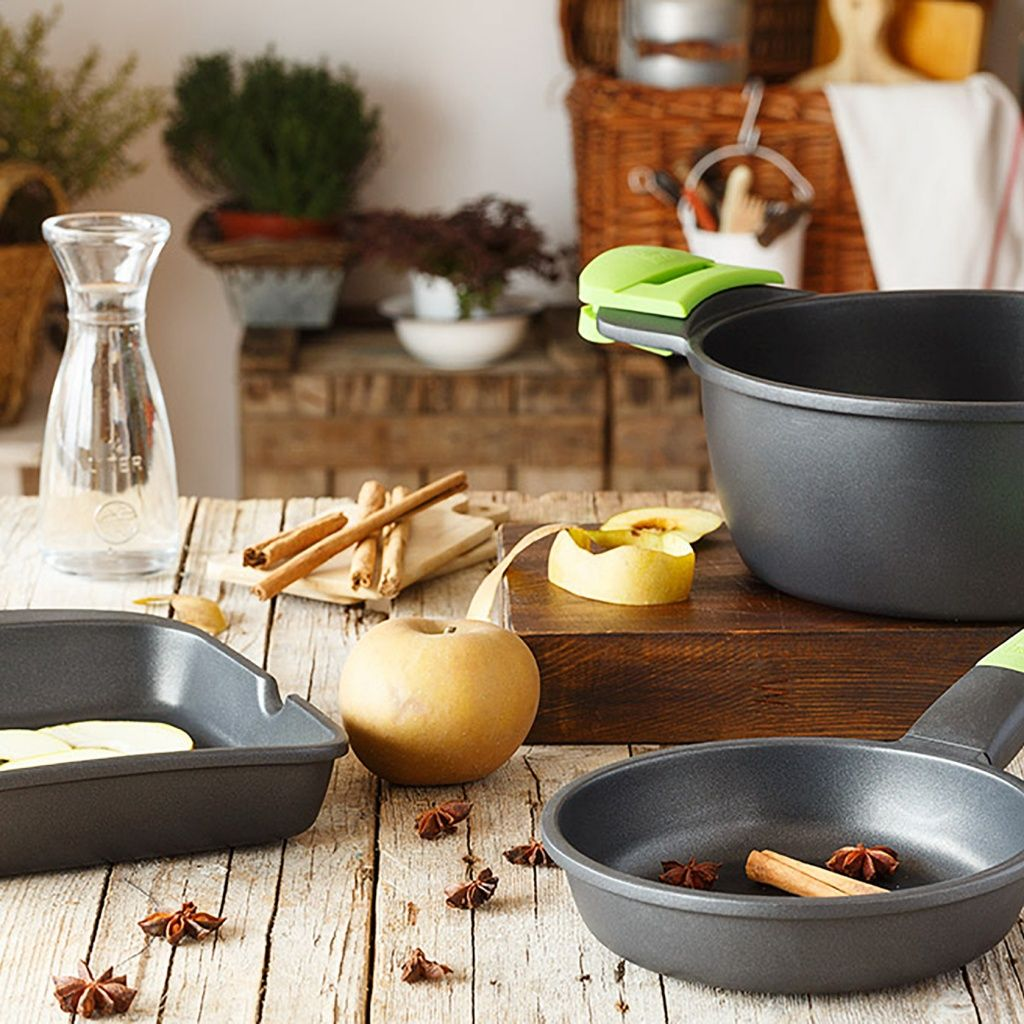 Una mesa de madera con utensilios de cocina, incluyendo sartenes, una manzana, palitos de canela y anís estrellado, con plantas detrás.