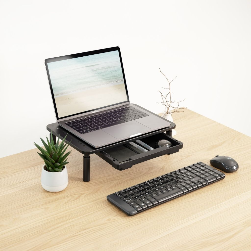 Laptop su supporto nero con cassetto. Tastiera, mouse, piccola pianta e rametti decorativi su scrivania in legno. Vista spiaggia sullo schermo.