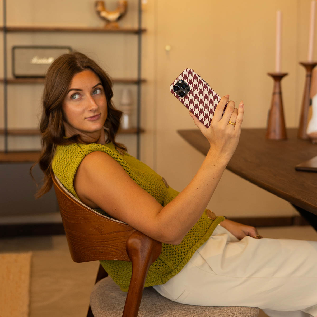 Vrouw in groene top maakt een selfie met een telefoonhoesje. Ze zit op een houten stoel in een kamer met planken en een tafel.