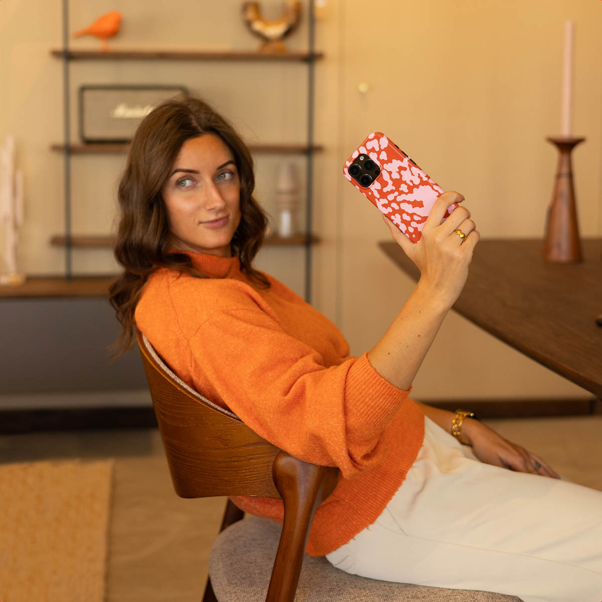 Een vrouw in een oranje trui houdt een telefoon met patroon vast. Ze zit op een houten stoel en kijkt in de camera. De achtergrond bevat planken.