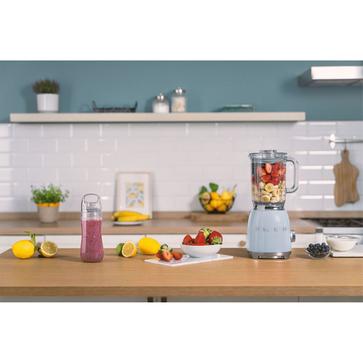 Scena kuchenna z blenderem wypełnionym owocami, butelką smoothie, cytrynami i truskawkami na drewnianym blacie.