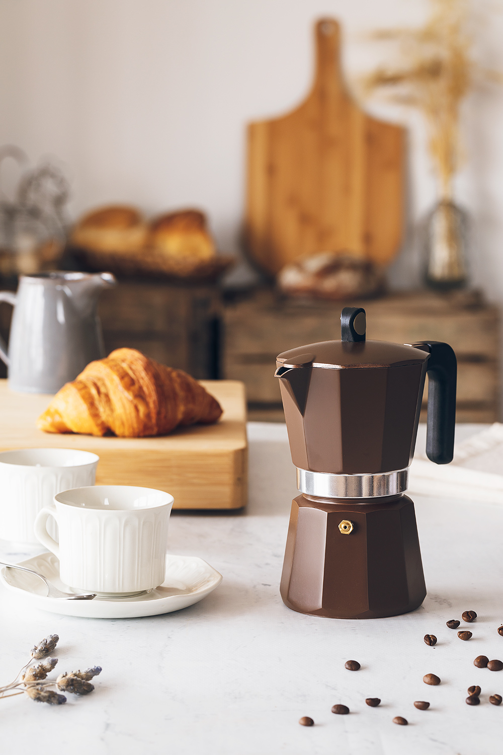 Brązowy ekspres do kawy, rogalik, filiżanki i ziarna kawy na stole. Inne przedmioty to chleb i deski do krojenia.