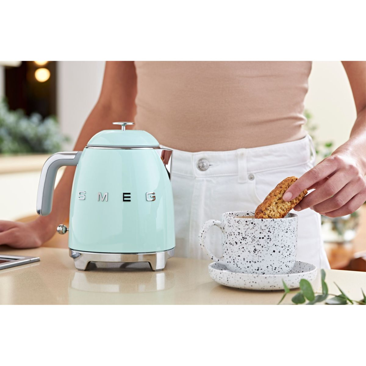 Kobieta macza biszkopt w nakrapianym kubku z kawą. Obok stoi miętowy czajnik. Kobieta ma na sobie beżowy top i białe spodnie.