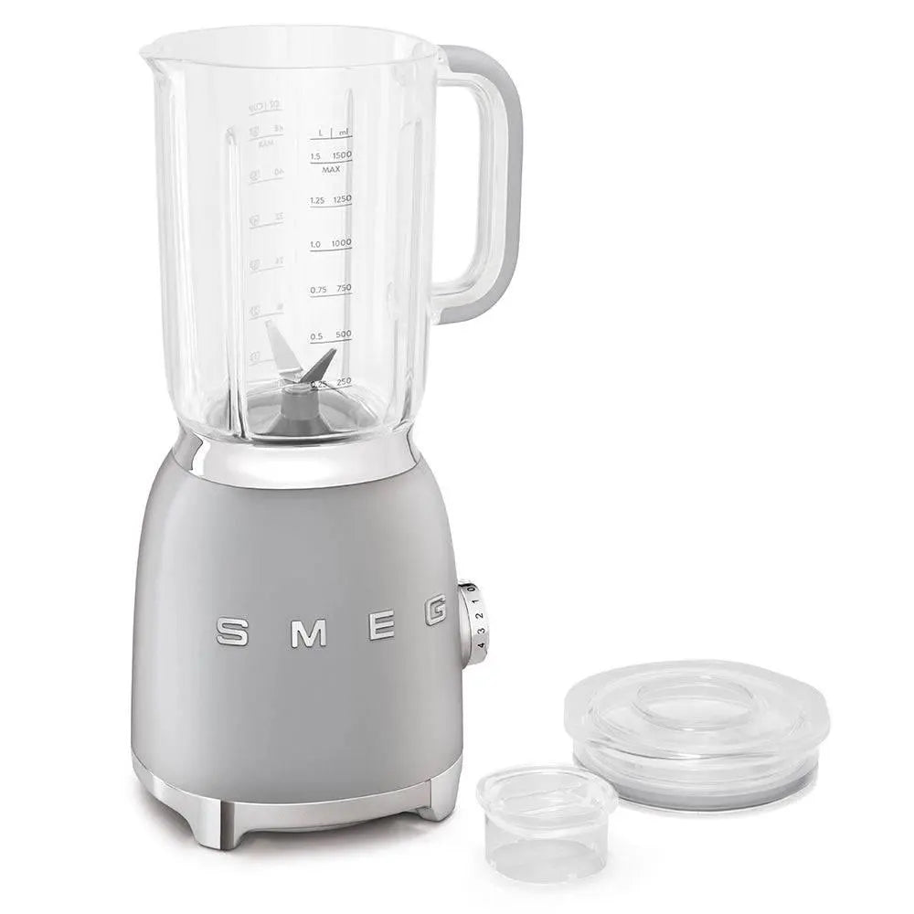 Una licuadora Smeg, base gris con una jarra transparente. Una tapa y una pequeña taza medidora están al lado. Los números están en la jarra.