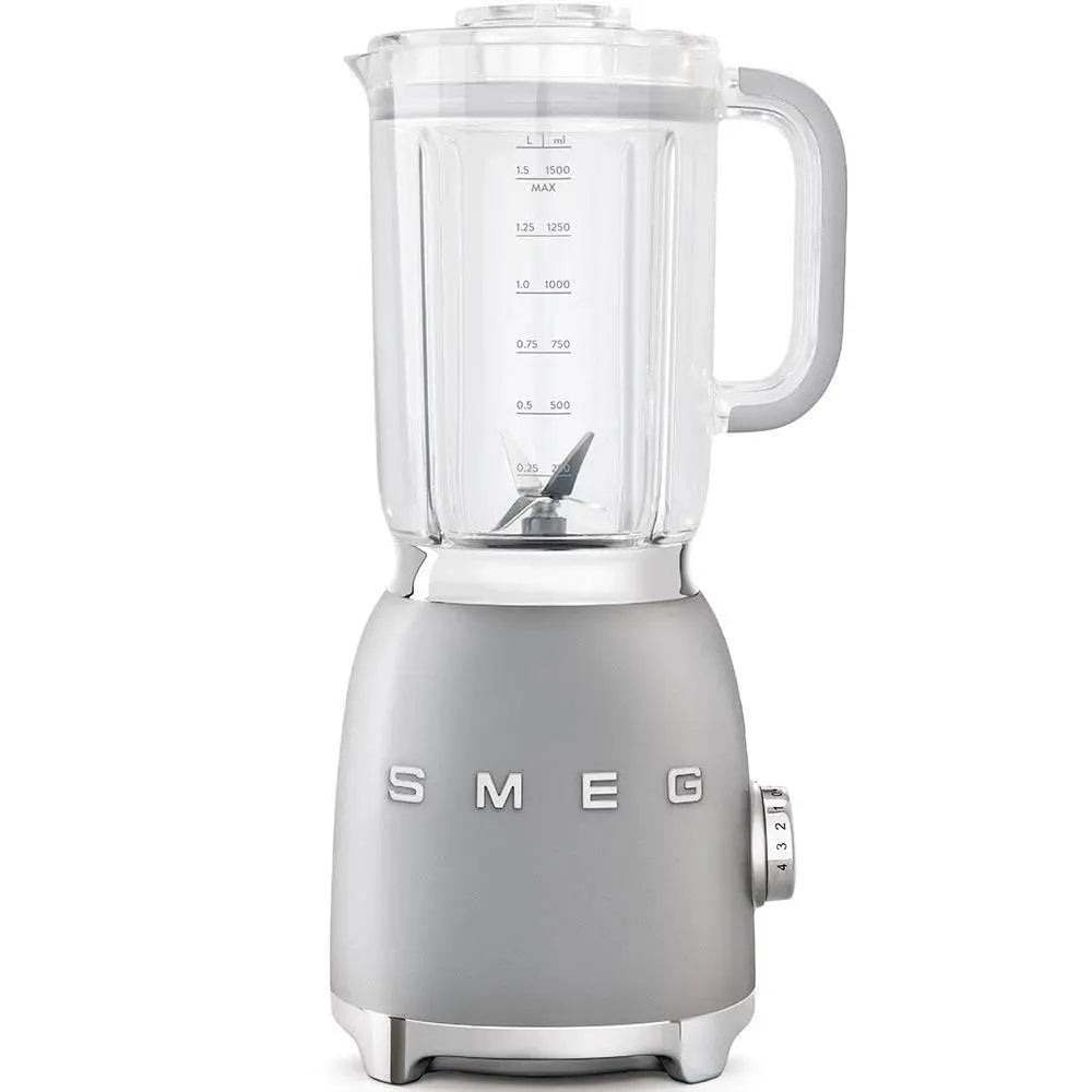 Una licuadora Smeg gris sobre fondo blanco. Tiene una jarra de vidrio y un dial. Marcas de volumen son visibles.