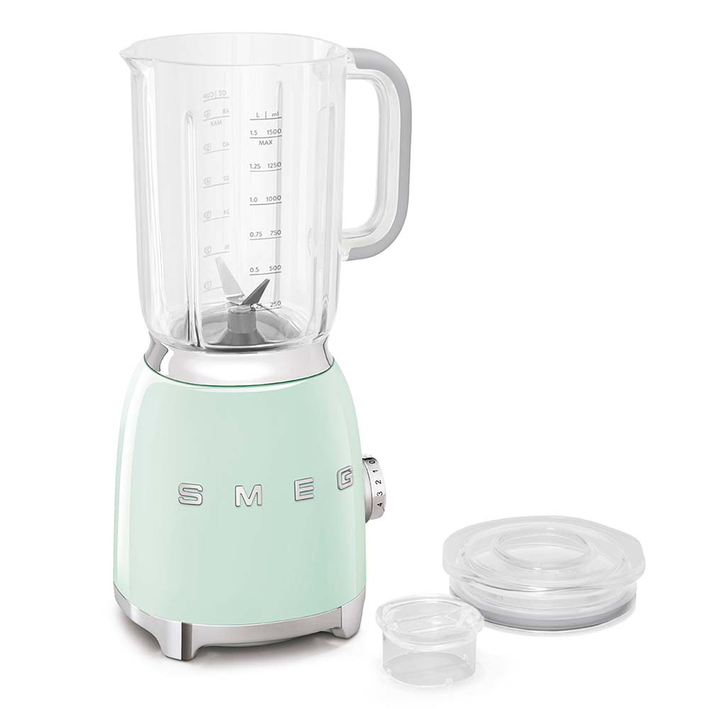 Un frullatore SMEG verde menta con un boccale di vetro trasparente, manico grigio e segni di misurazione. Due coperchi di plastica trasparente accanto.