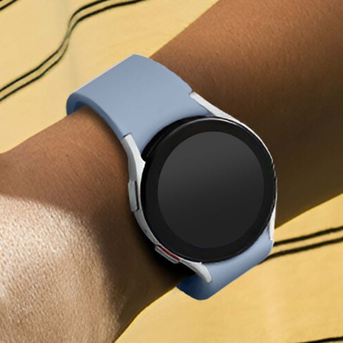Un polso con uno smartwatch azzurro. L'orologio ha uno schermo nero e una cornice argentata. Lo sfondo è giallo con strisce nere.