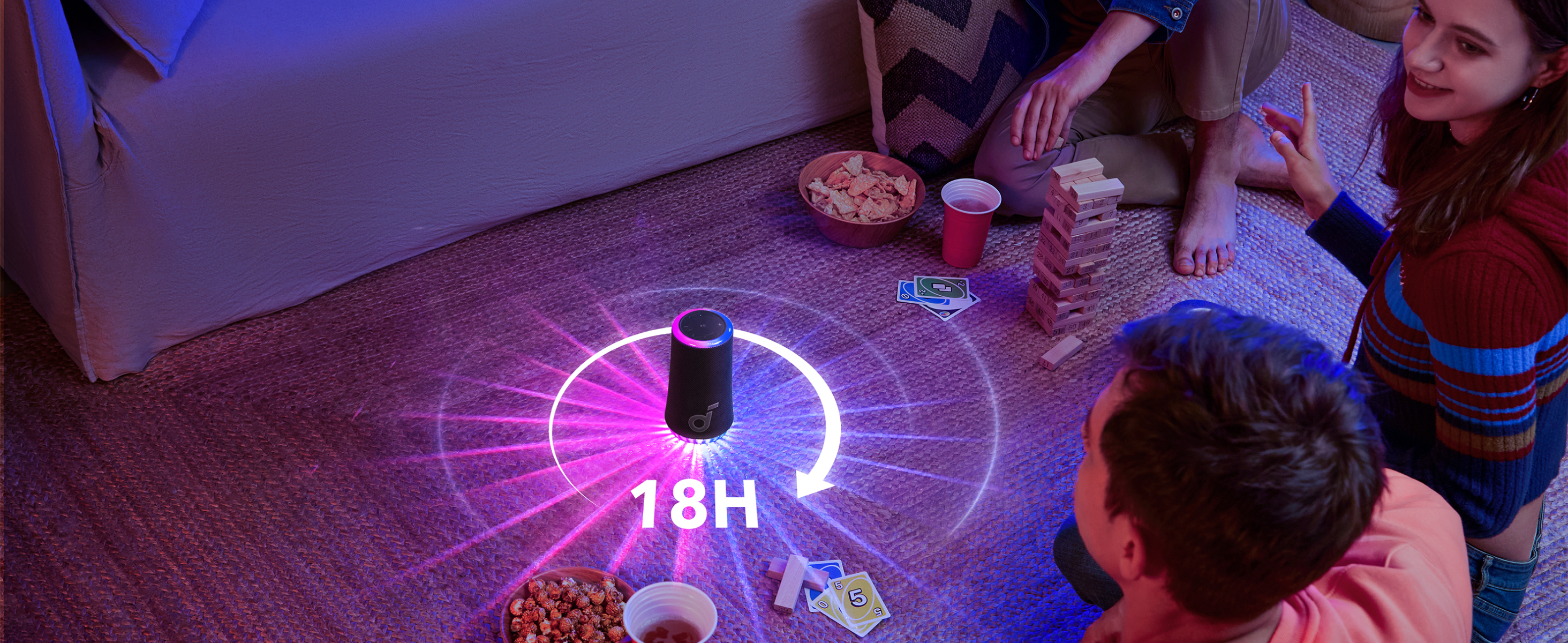 Przyjaciele grają w gry wokół głośnika z wyświetlaczem 18H. Widoczne przekąski, napoje i wieża Jenga.