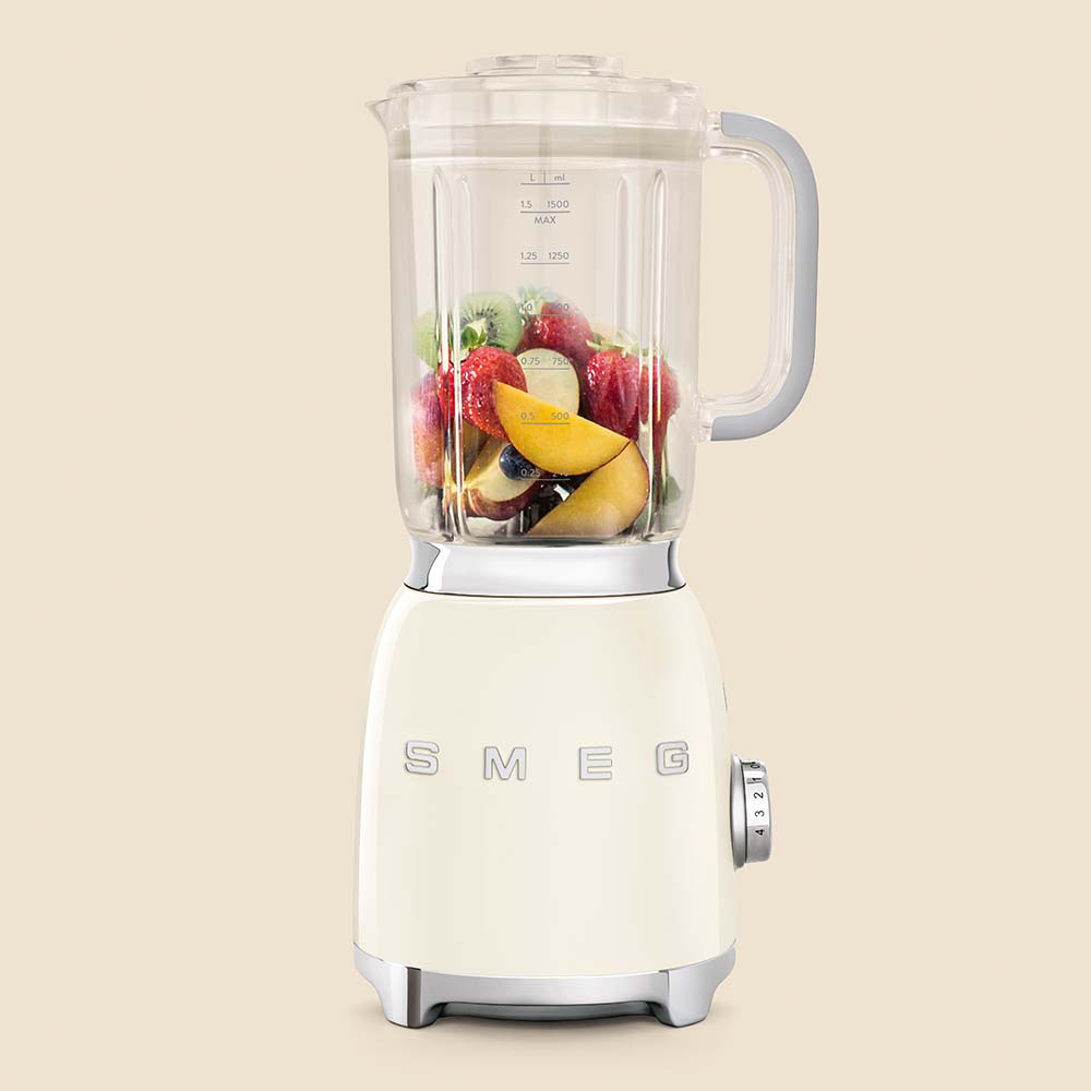 Licuadora SMEG color crema con fruta dentro. Se ven fresas, kiwi y rodajas de melocotón. Fondo beige.