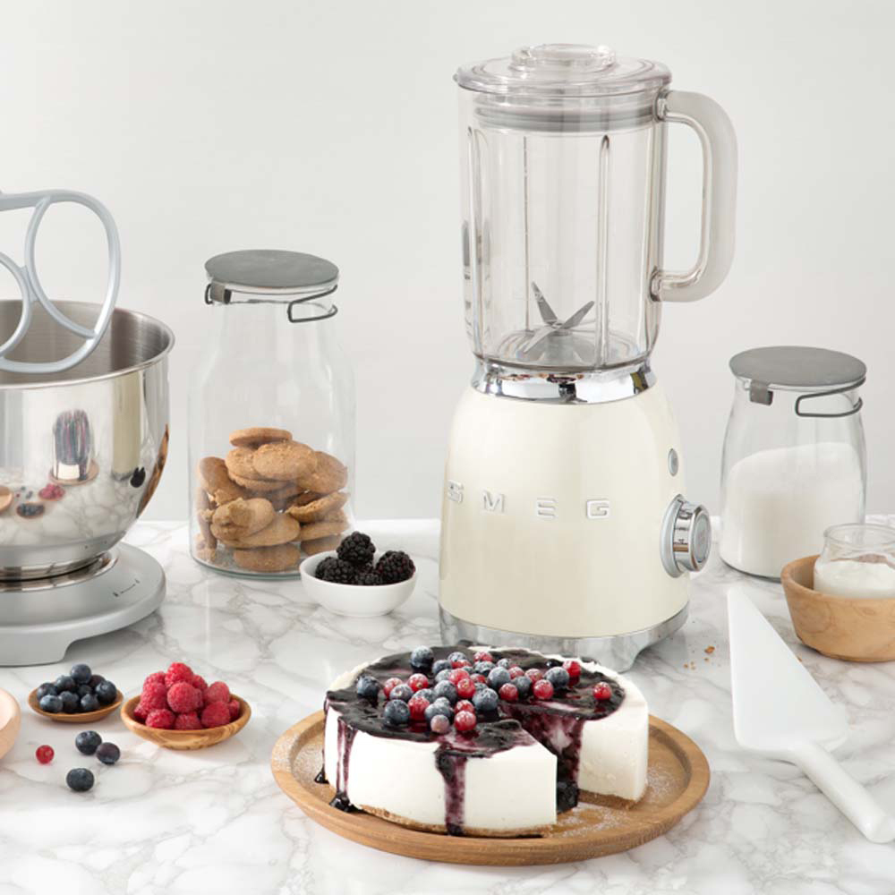 Una escena de cocina con licuadora, pastel, bayas, galletas y batidora. El pastel está cubierto con bayas y salsa. Otros ingredientes son visibles.