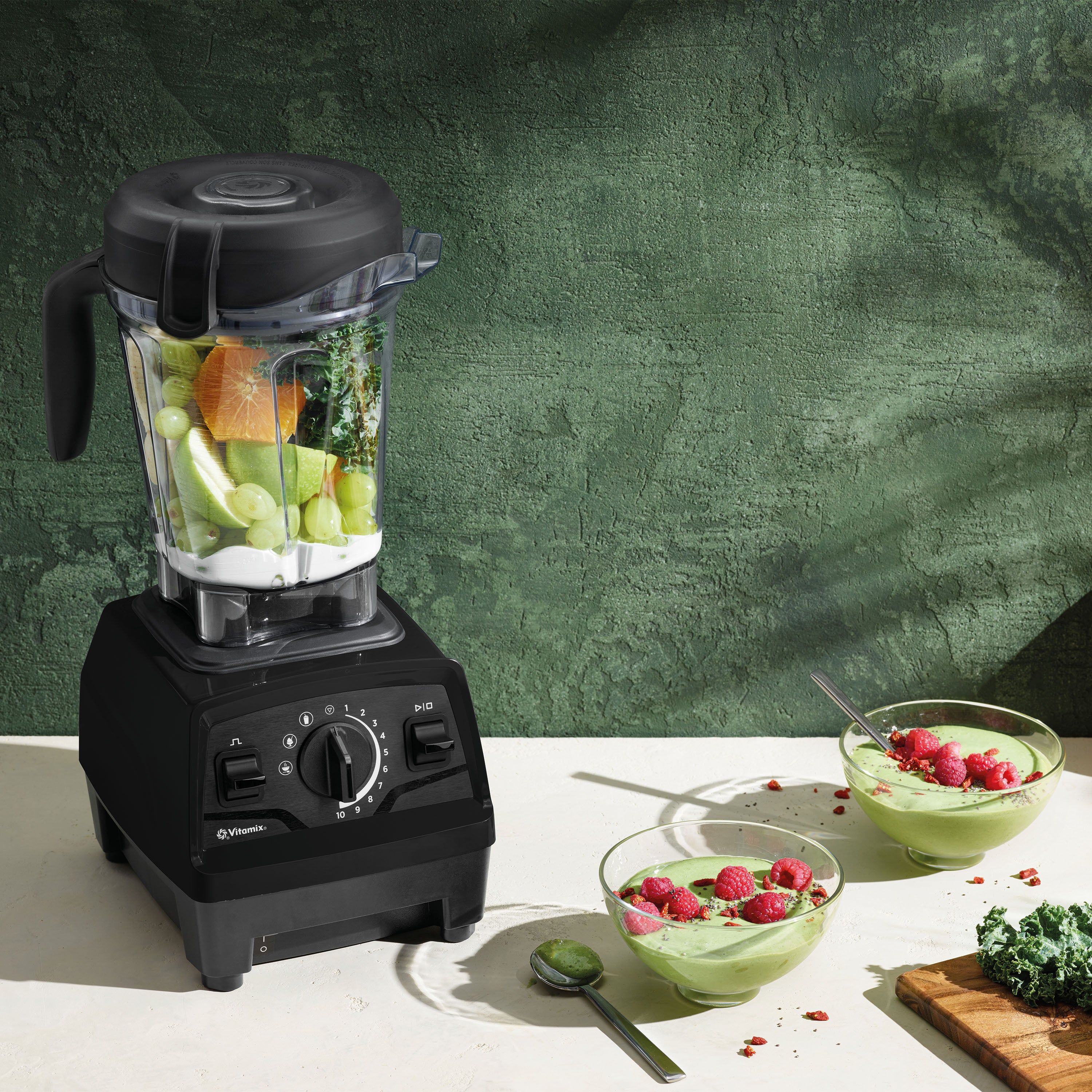 Ein schwarzer Mixer mit Früchten. Zwei Schalen mit grünem Smoothie und Himbeeren auf weißer Oberfläche.