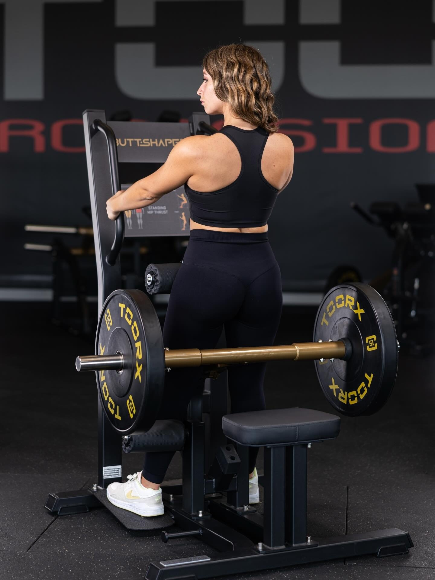 Een vrouw in zwarte sportkleding gebruikt een gewichtmachine. De machine heeft aan beide kanten gewichten en de woorden 'BUTT-SHAPE' zijn zichtbaar.