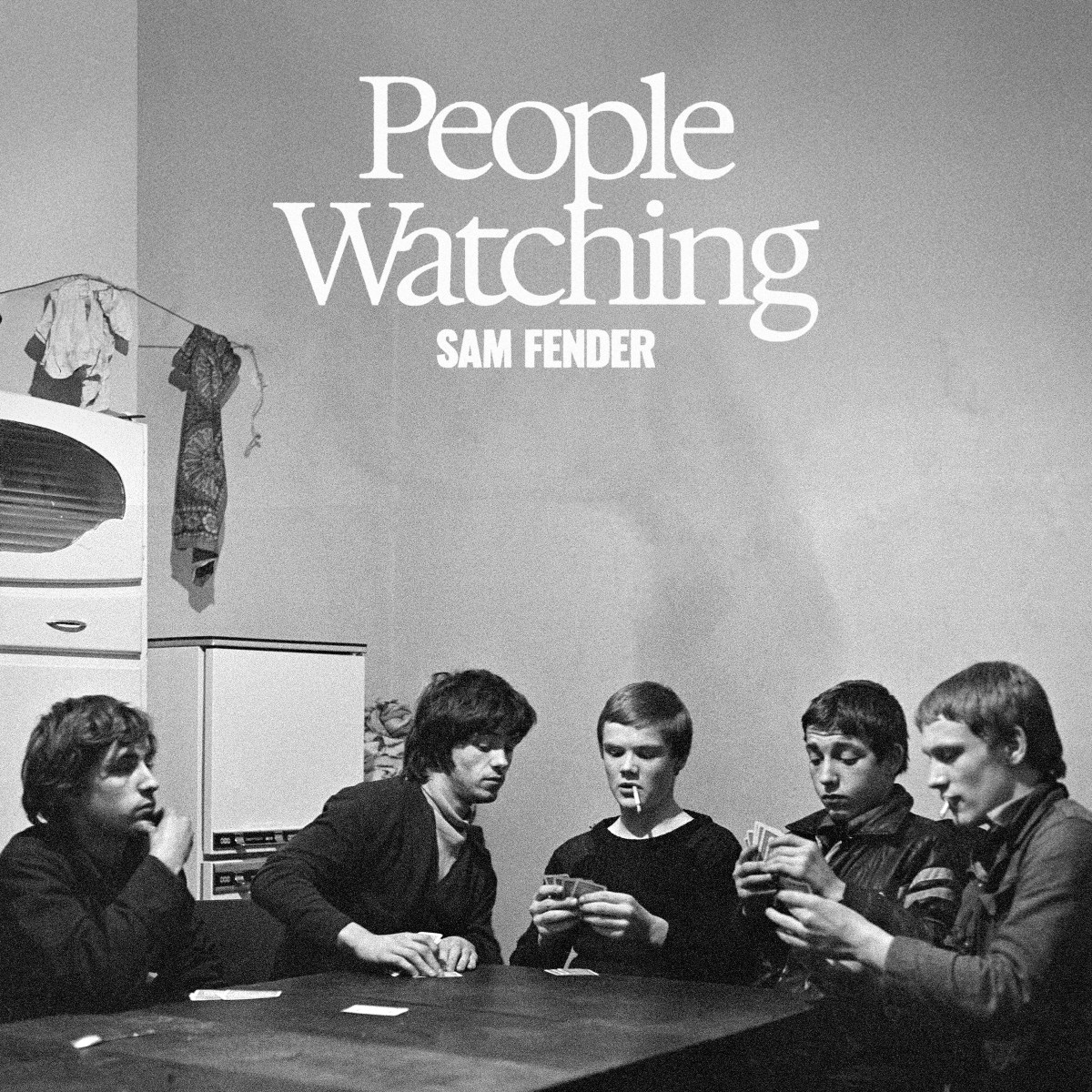 Cinque uomini seduti attorno a un tavolo giocano a carte. Sigarette in bocca. Le parole "People Watching" e "Sam Fender" sono sopra.