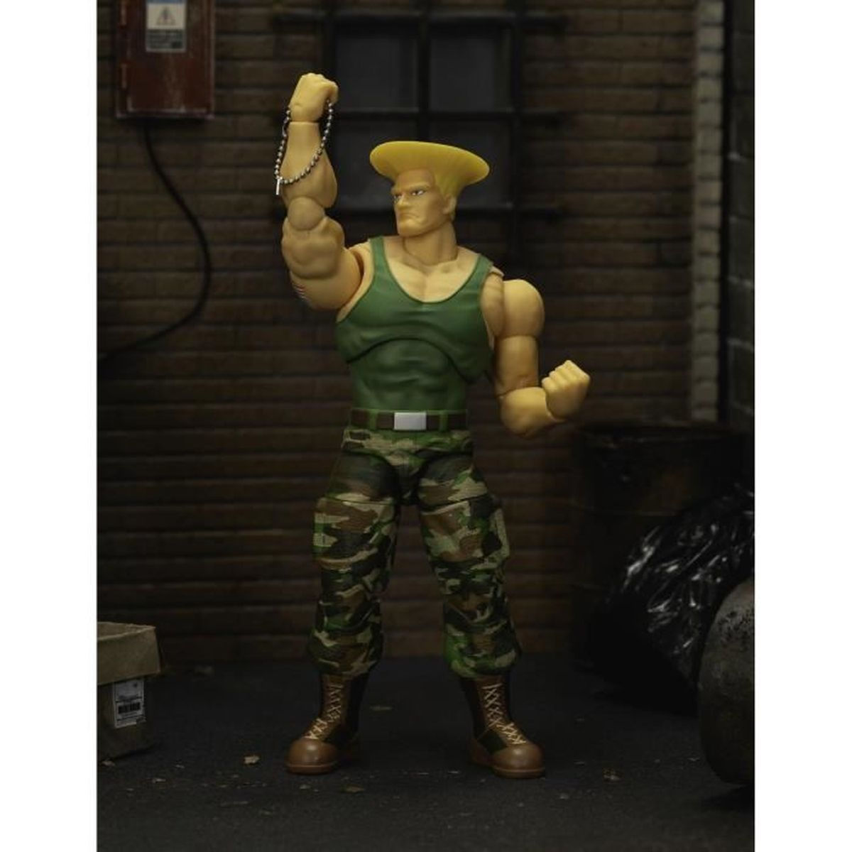 Eine Figur von Guile aus Street Fighter. Er trägt ein grünes Tanktop, Camouflage-Hose und Stiefel. Er hebt den Arm zu einer Faust mit einer Kette.