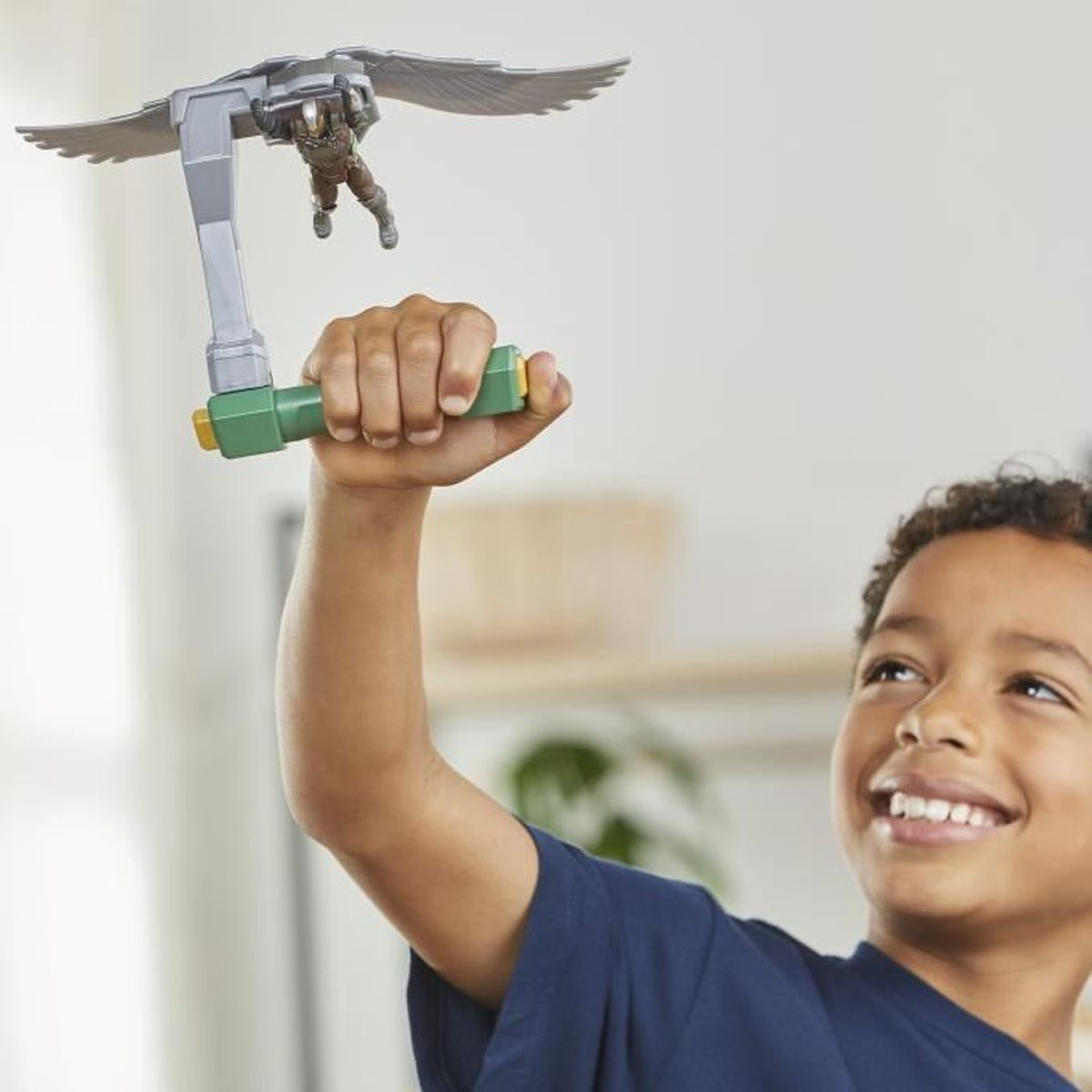 Ein Junge hält ein Spielzeug mit einer Figur und Flügeln. Er lächelt nach oben. Das Spielzeug hat einen grünen Griff und graue Flügel.