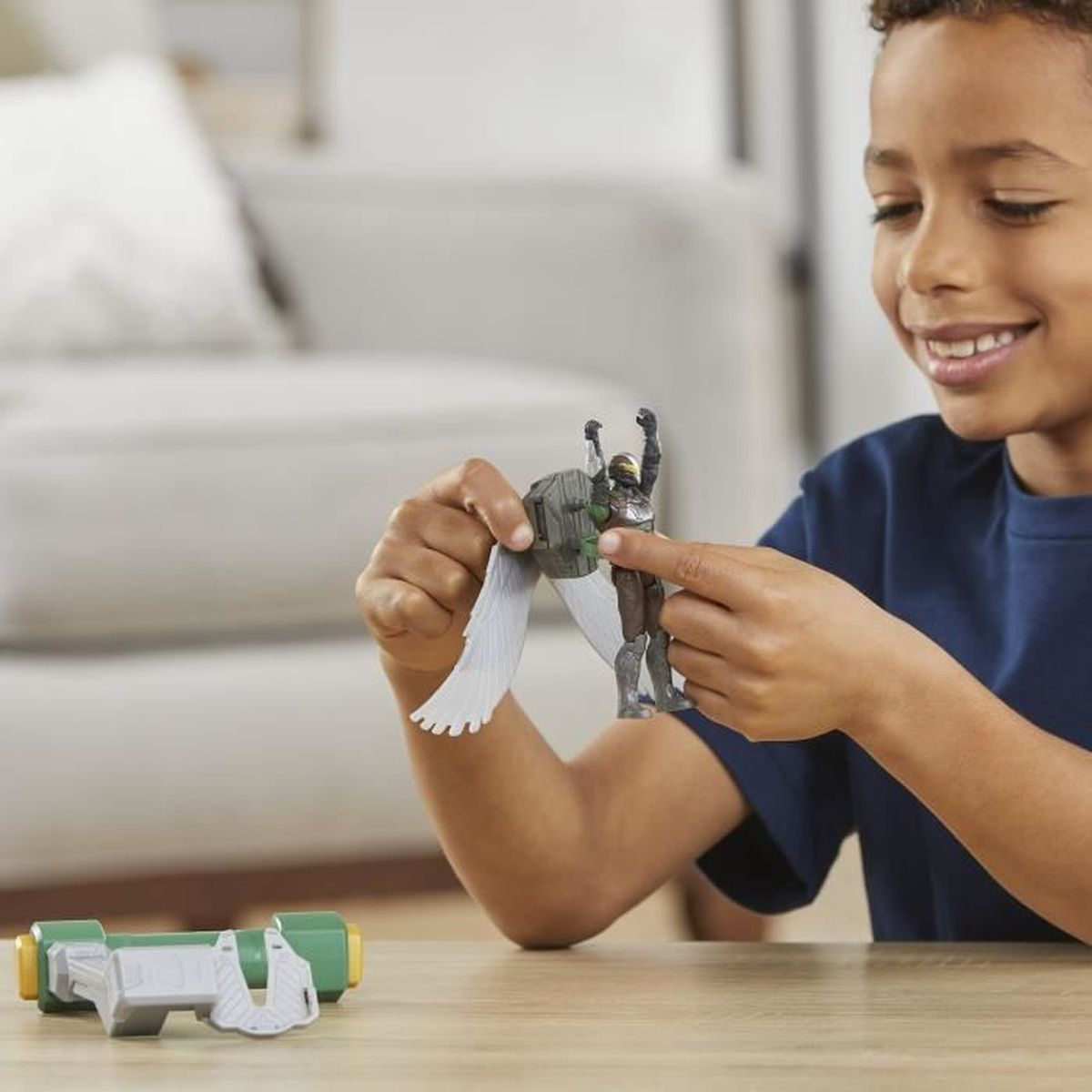Ein Junge lächelt, während er eine Spielzeug-Actionfigur mit Flügeln hält. Ein grünes und graues Spielzeug befindet sich im Vordergrund.