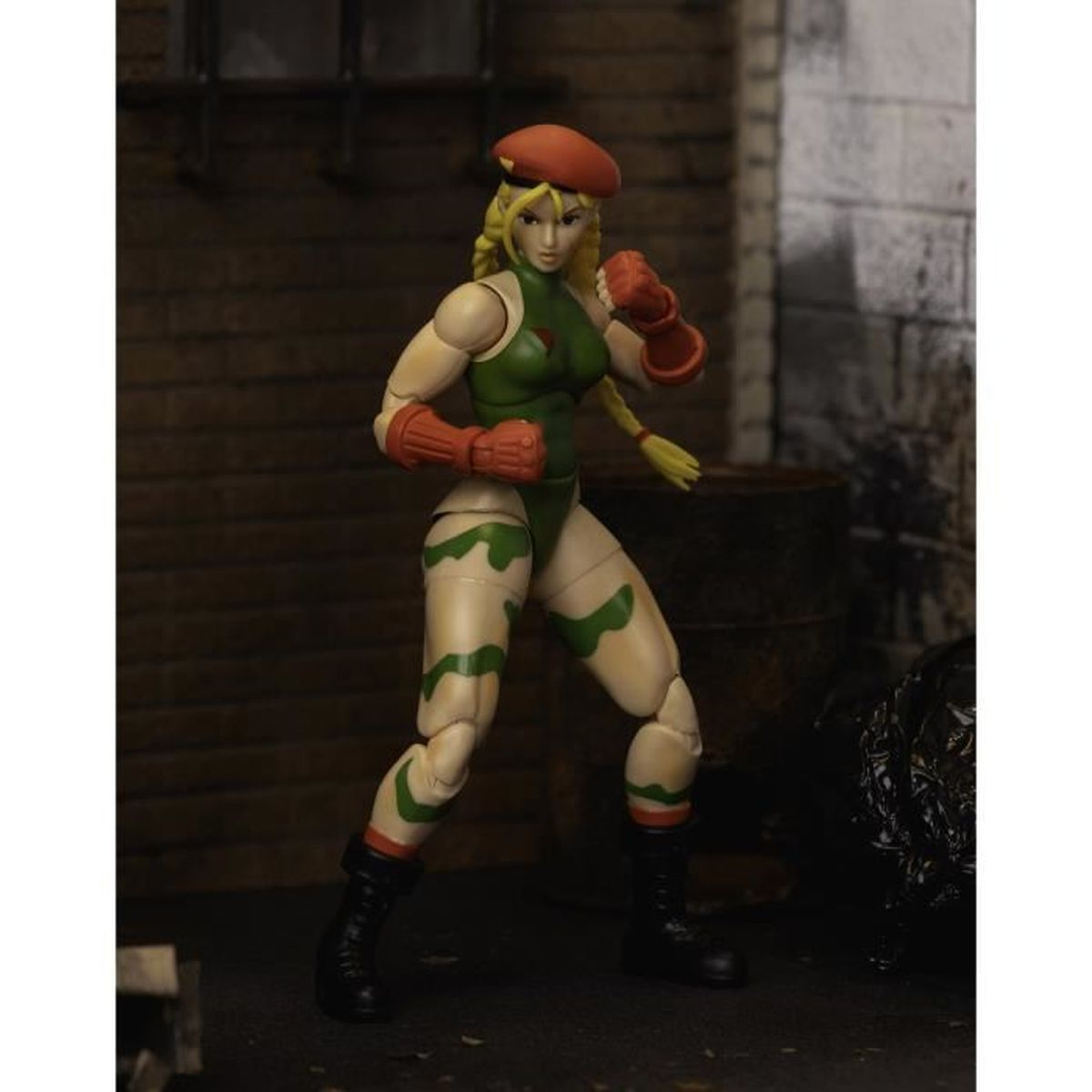 Eine Cammy-Actionfigur steht bereit. Sie trägt einen grünen Body, orangefarbene Handschuhe und eine Baskenmütze. Ihr blondes Haar ist geflochten. Hinter ihr steht ein Fass.