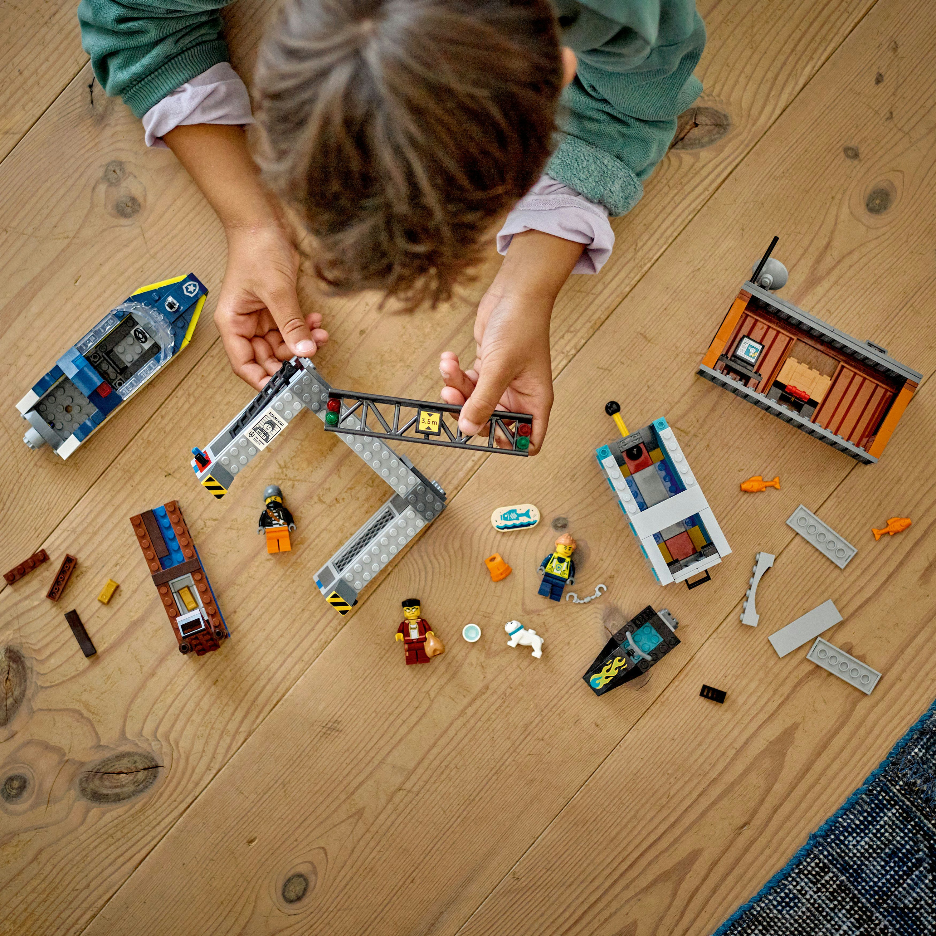 Dziecko bawiące się policyjną łodzią Lego, figurkami i elementami konstrukcyjnymi na drewnianej podłodze. Widoczne dłonie.