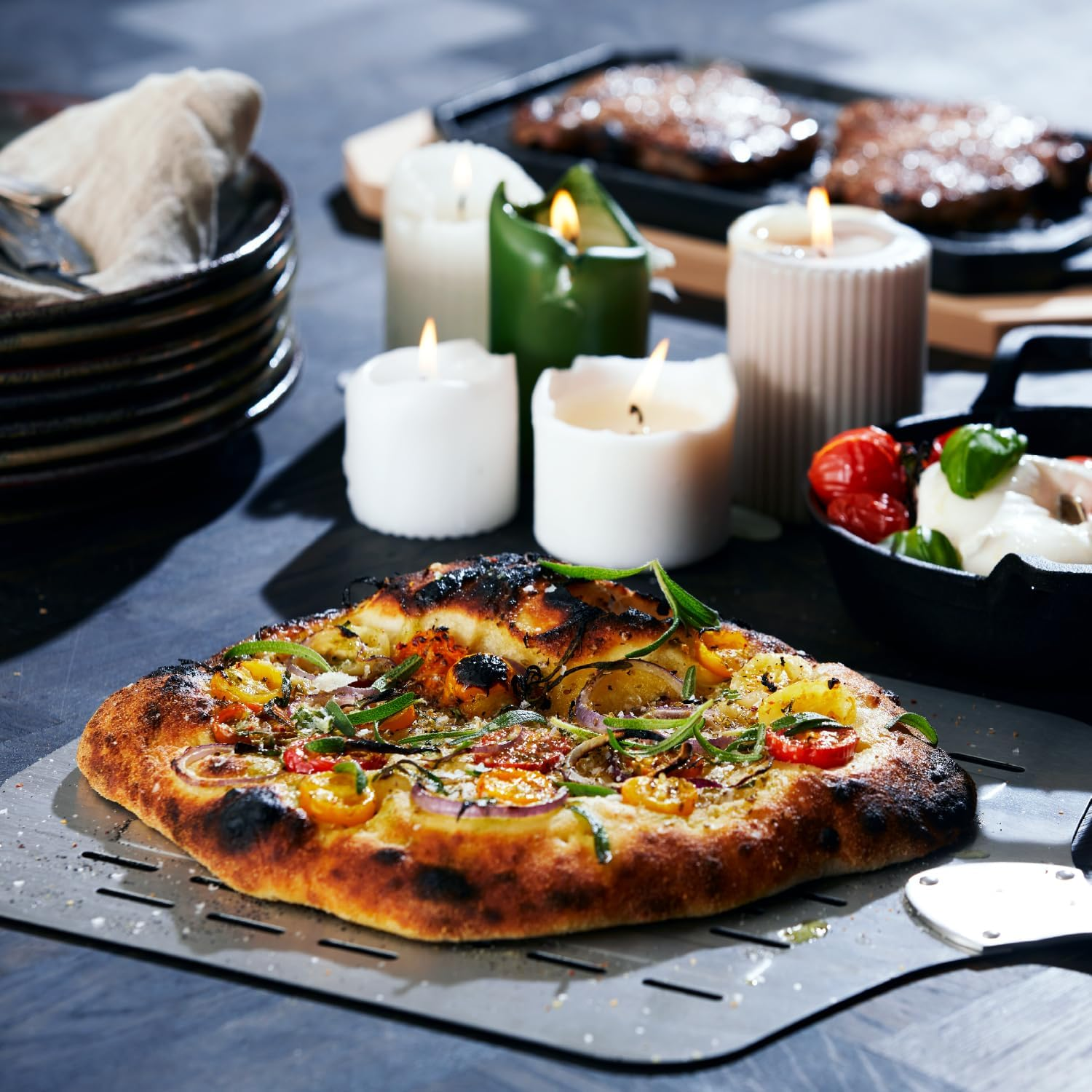 Pizza auf einer Metallplatte, mit Kerzen und Essen im Hintergrund. Pizza hat Gemüse. Dunkle Umgebung.