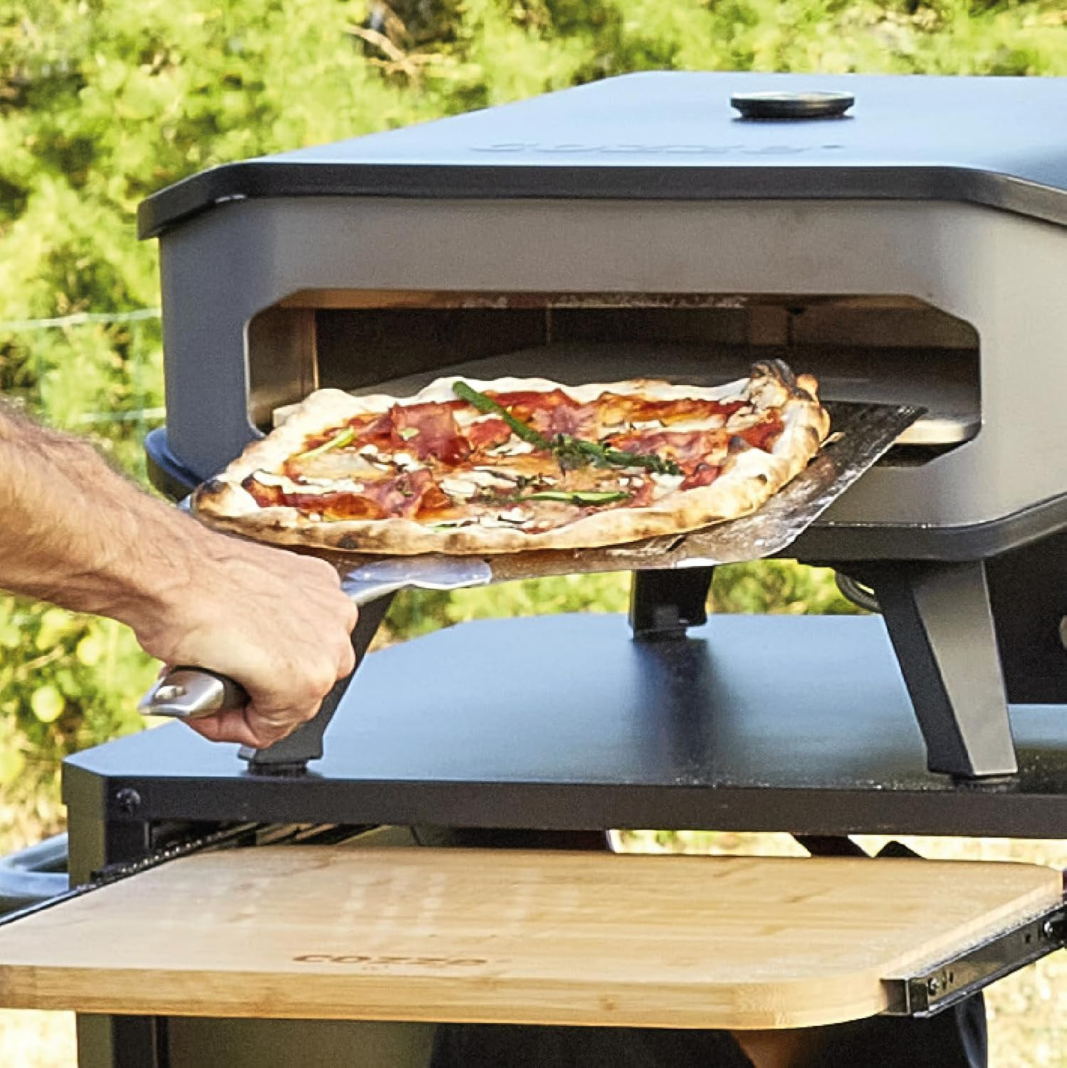 Eine Person nimmt mit einem Pizzaschieber eine Pizza aus einem Pizzaofen. Die Pizza hat Belag. Unten ist ein Holzbrett.