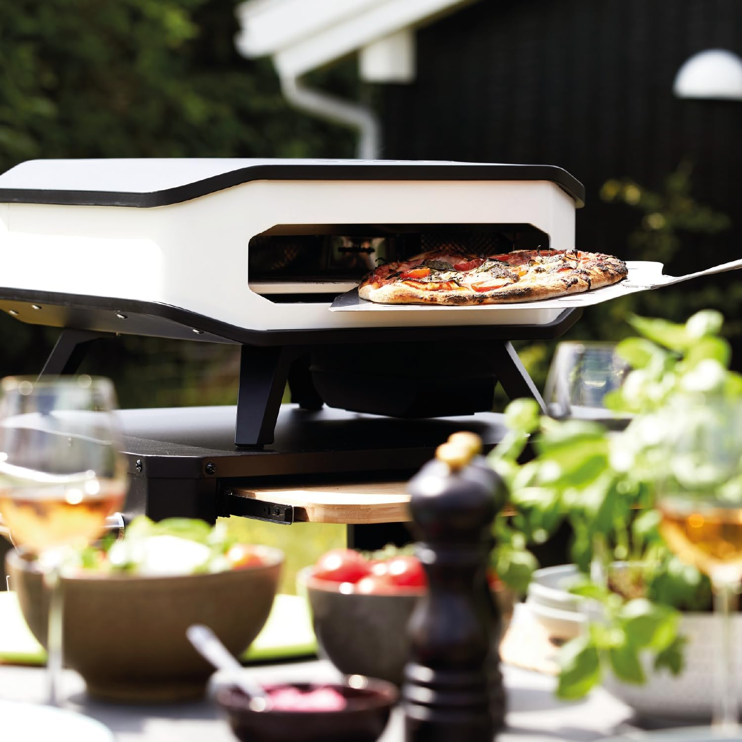 Pizza wird aus einem Pizzaofen im Freien genommen. Ein Tisch ist mit Essen, Weingläsern und Grünzeug in einem Garten gedeckt.