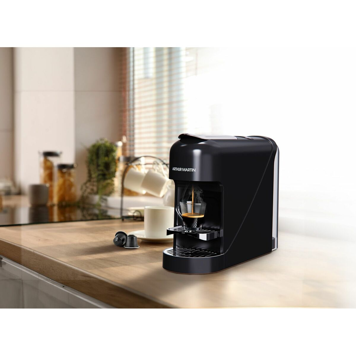 Una macchina da caffè Arthur Martin nera che prepara il caffè in una tazza di vetro. Una tazza e capsule sono su un bancone di legno. Lo sfondo è una cucina.