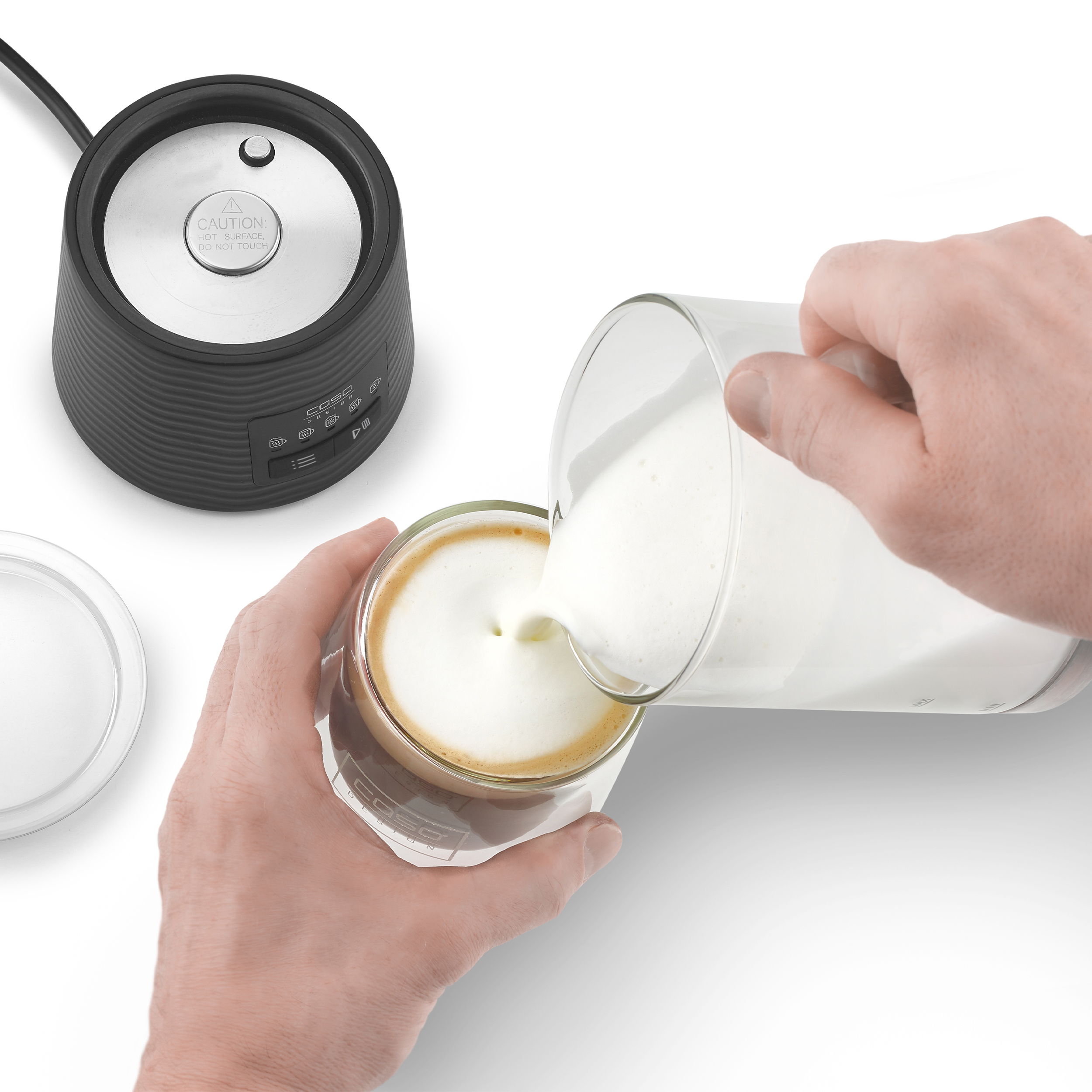 Hände gießen Milchschaum in ein Kaffeeglas. Links ist ein Milchaufschäumer.