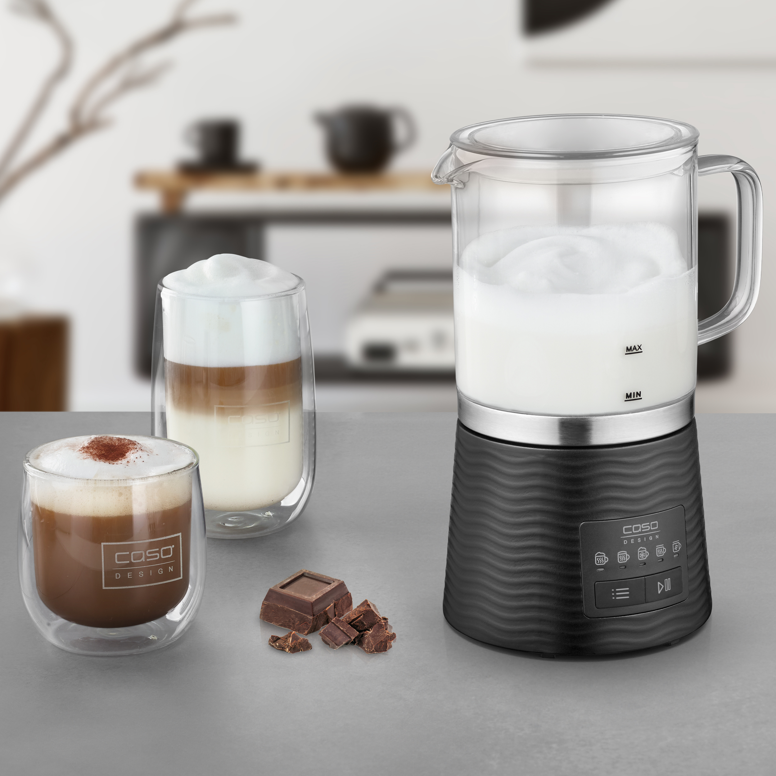 Ein Milchaufschäumer, zwei Kaffeegläser und Schokoladenstücke auf einer grauen Oberfläche. Verschwommener Hintergrund zeigt eine Küche.