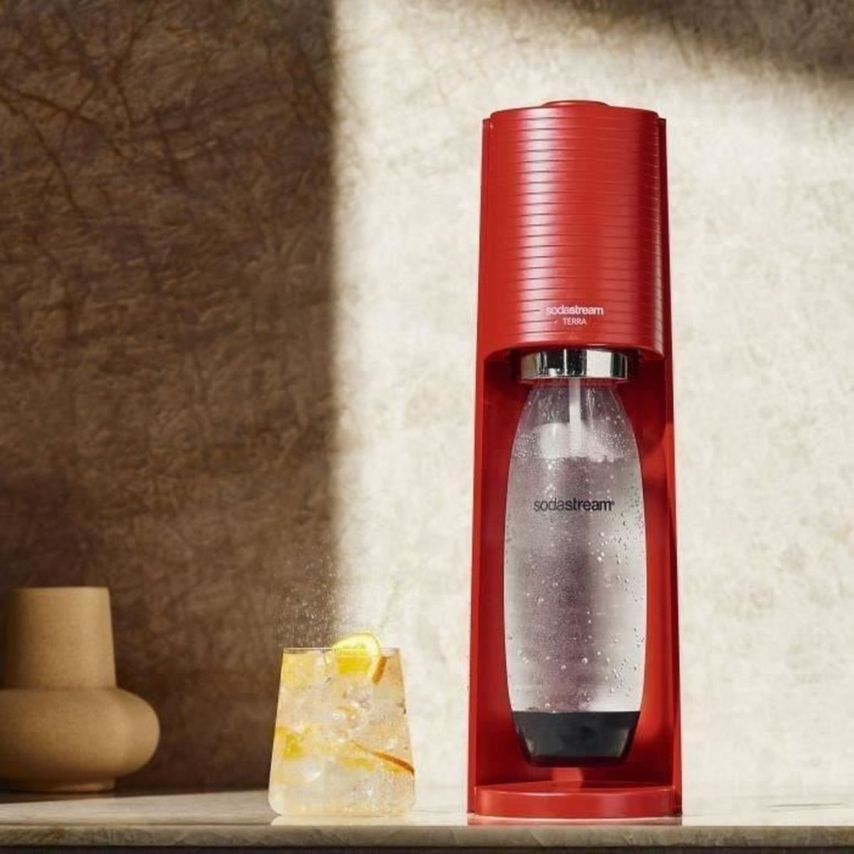 Rote SodaStream-Maschine mit einem Glas Cocktail mit Zitronenscheiben. Links steht eine Vase.